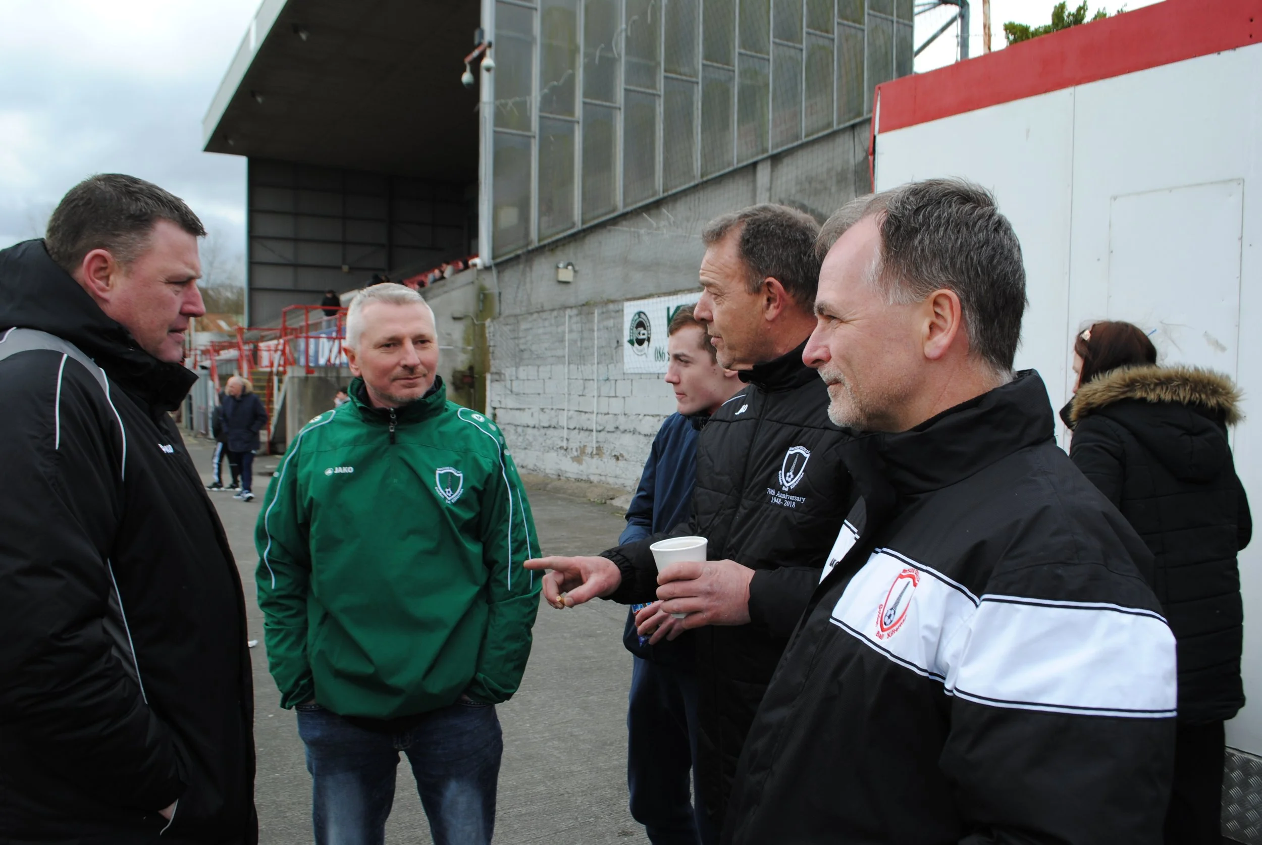 A group of men and women standing outside near a sports stadium having a conversation. One man is handing a paper cup to another man.