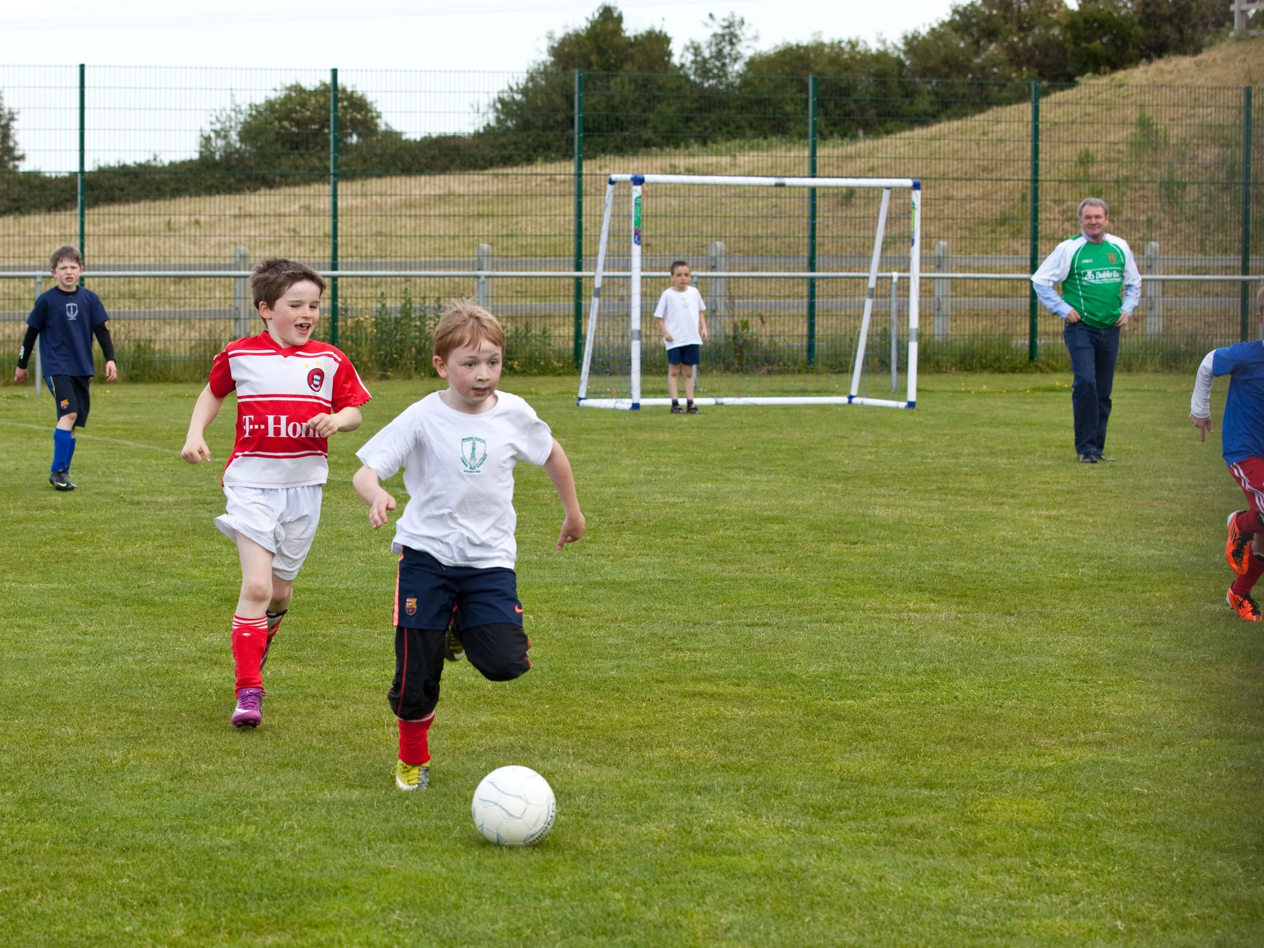 Children playing soccer on a grassy field with an adult supervising. Kids are running towards a white soccer ball, some wearing white and others wearing team jerseys. There is a soccer goal in the background, along with a fence and trees on a cloudy 