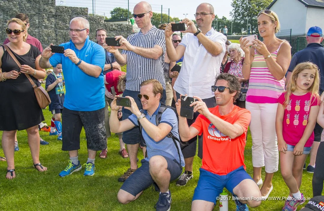 Group of people taking photos at an outdoor event on a sunny day, standing or kneeling on grass, with children and a fence in the background.
