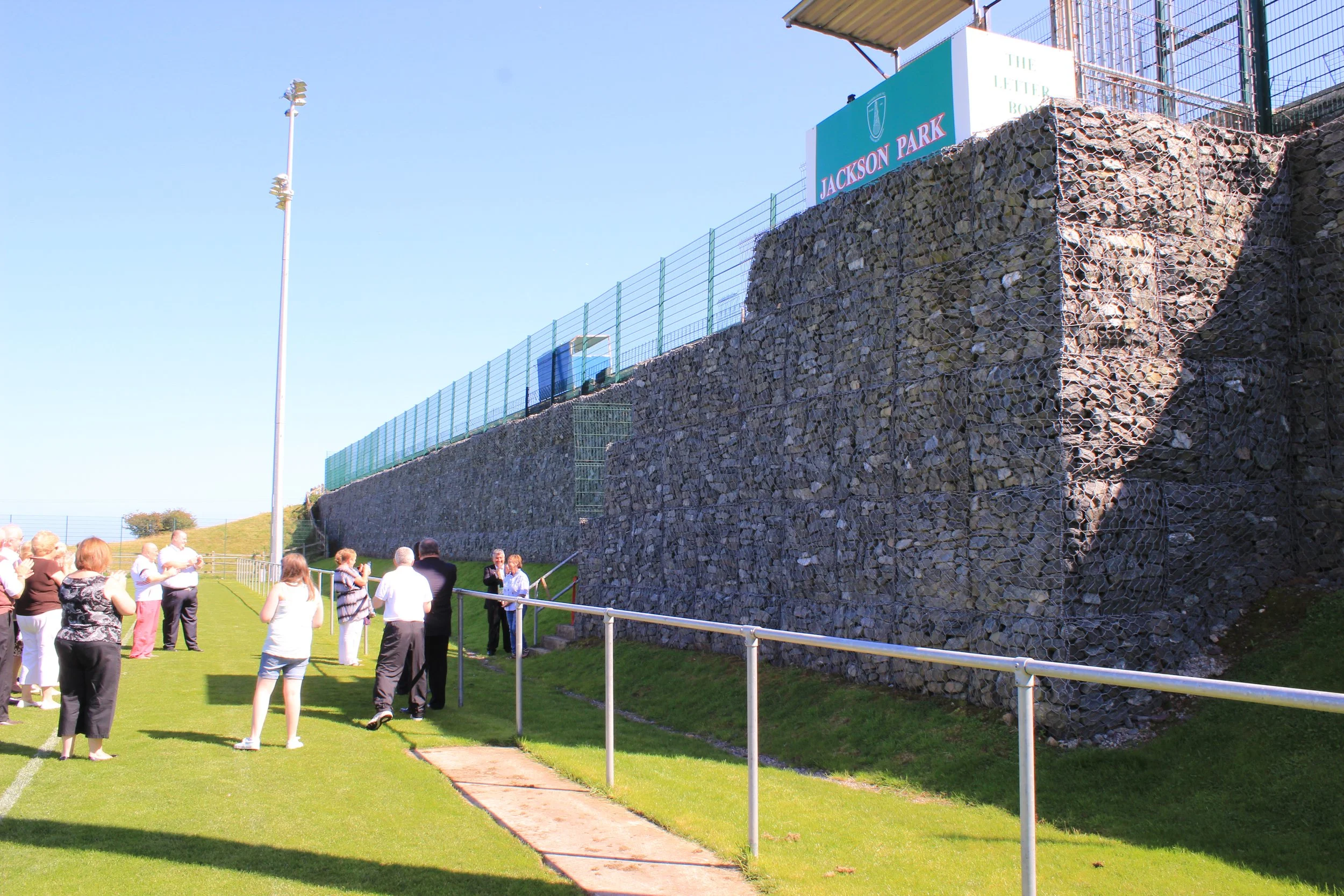 People gathered on a grassy field near a large stone wall with a sign that reads 'Jackson Park' and a small track with a metal fence surrounding it.