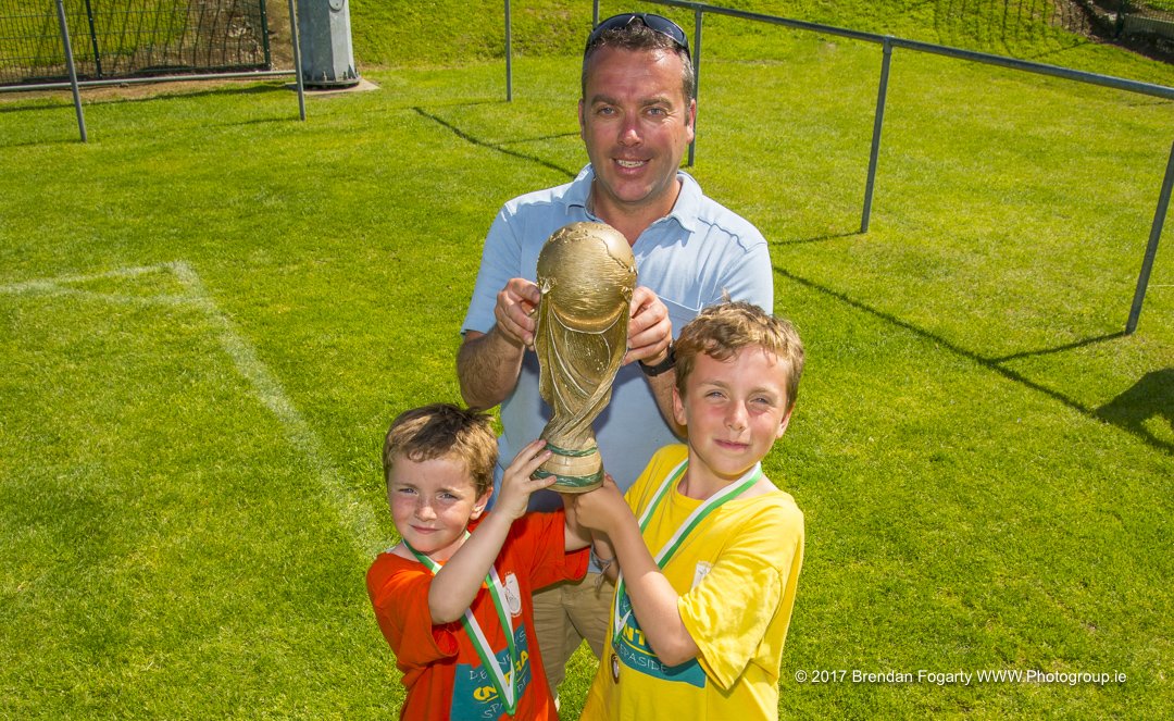 A man and two young boys standing on a grassy field, holding a large trophy together after a sports game or tournament, smiling at the camera.