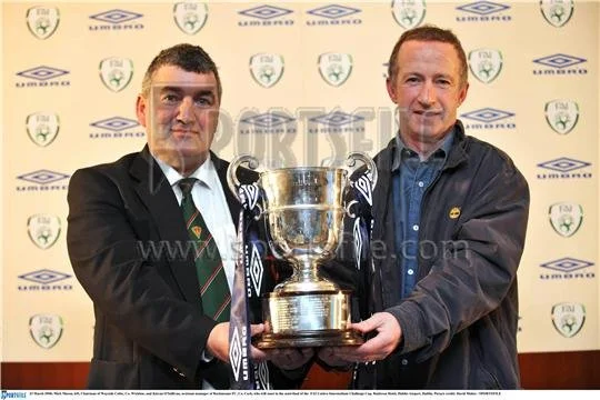 Two men in formal and casual attire holding a large trophy together, standing in front of a backdrop with sports logos.