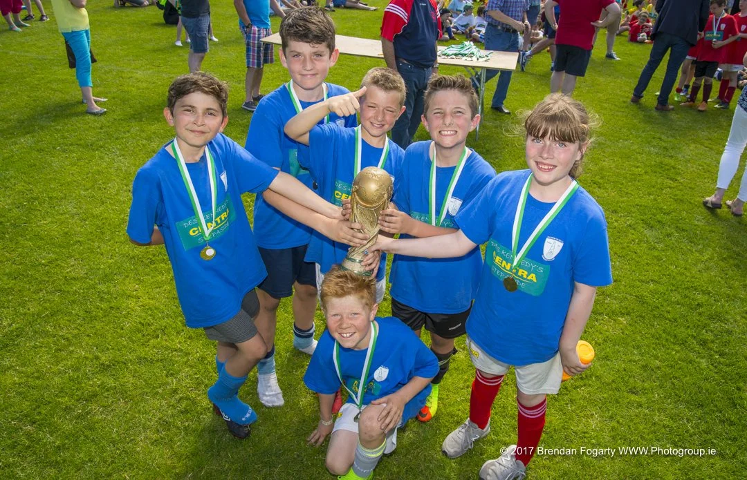A group of young children wearing blue shirts and medals, holding a large trophy, celebrate on a grassy field after a sports event.