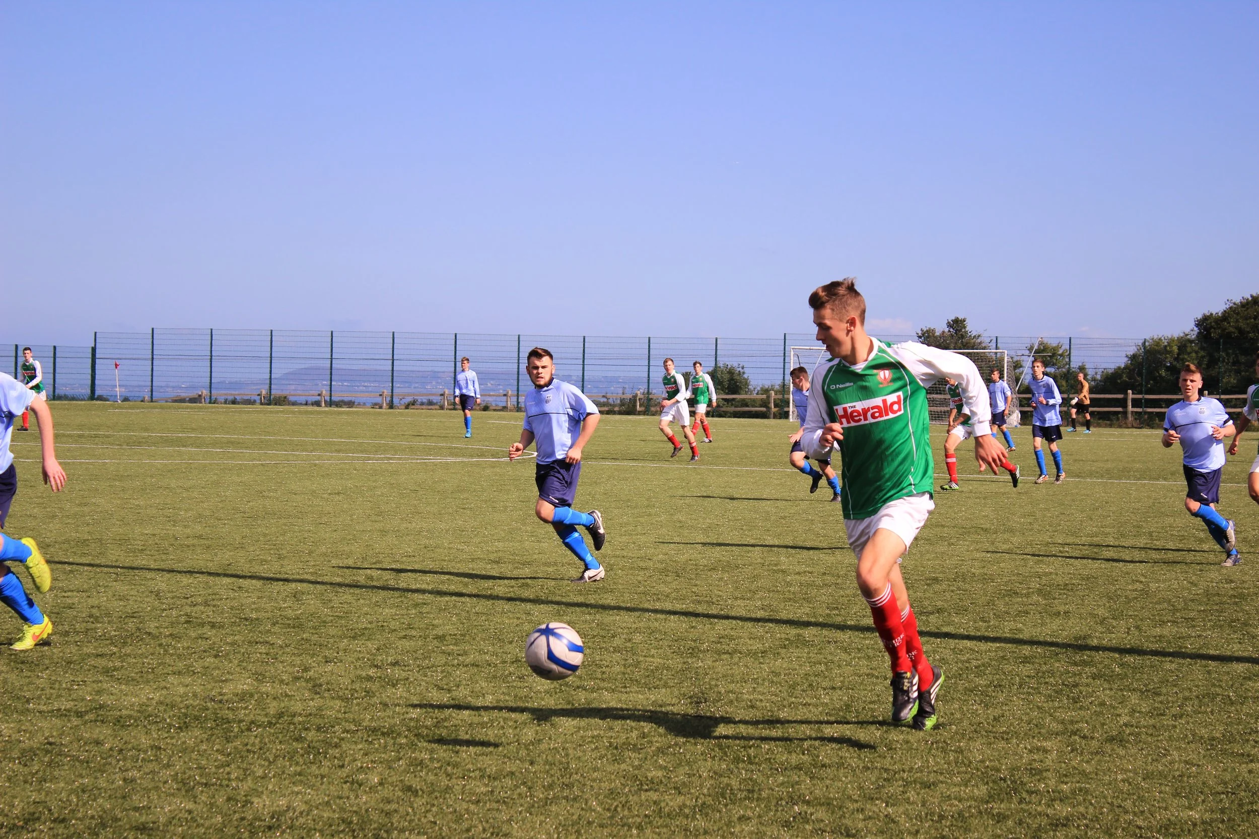 Young male soccer player in green and white uniform chasing the ball during a match on a grassy field under a clear blue sky, with other players and a fence in the background.