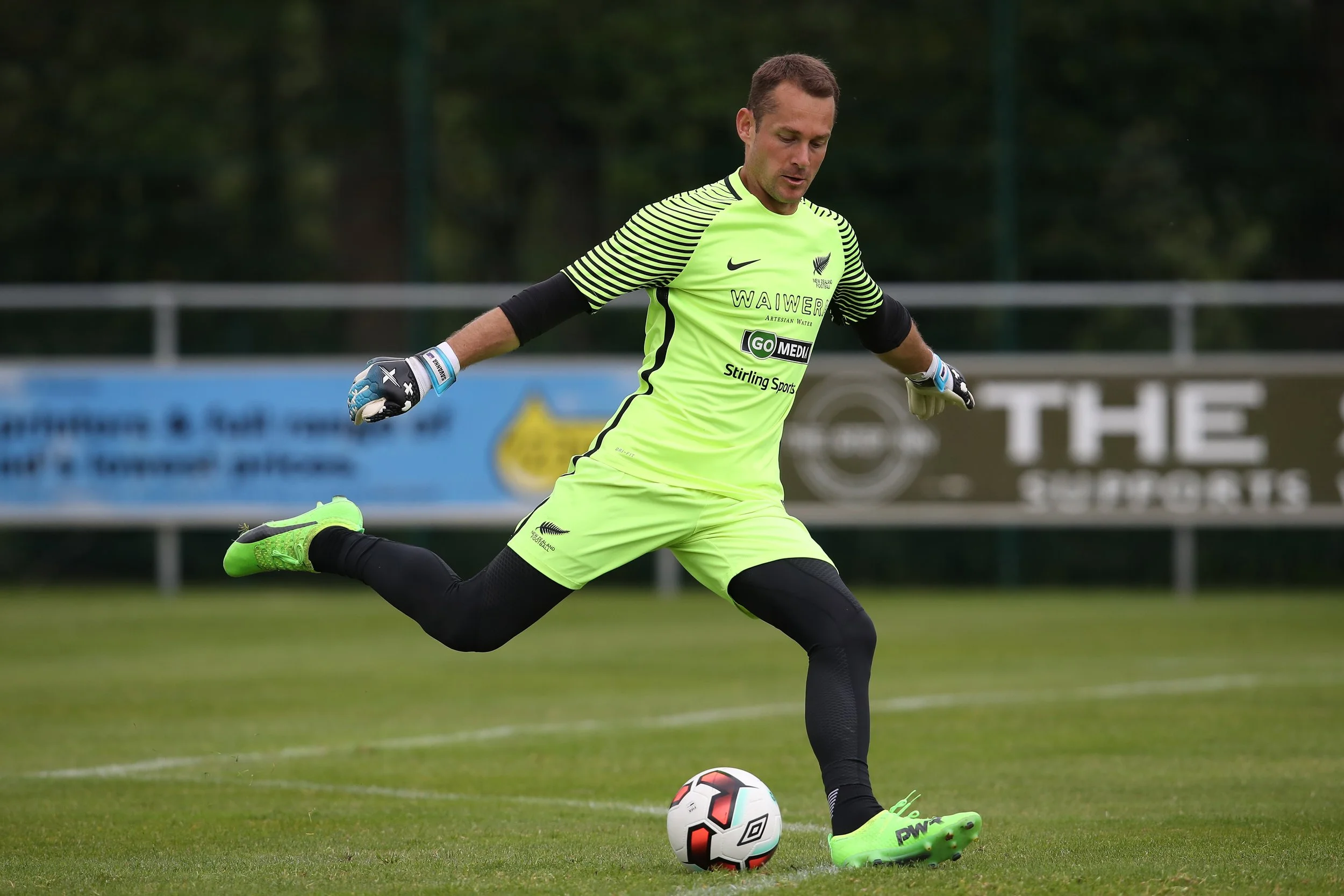 A male soccer player wearing a neon yellow uniform with black accents and goalkeeper gloves is kicking a soccer ball on a grass field during daytime. Background includes a fence and trees.