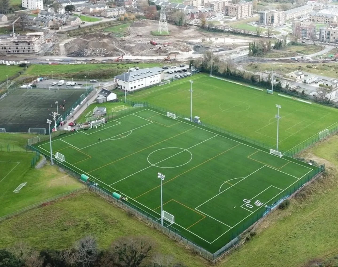 Aerial view of two adjacent soccer fields, one with artificial turf and the other with natural grass, in a sports complex surrounded by fences, with light poles and a small building nearby. In the background, there is construction site with dirt, equipment, and partially built structures, as well as residential houses and city buildings.