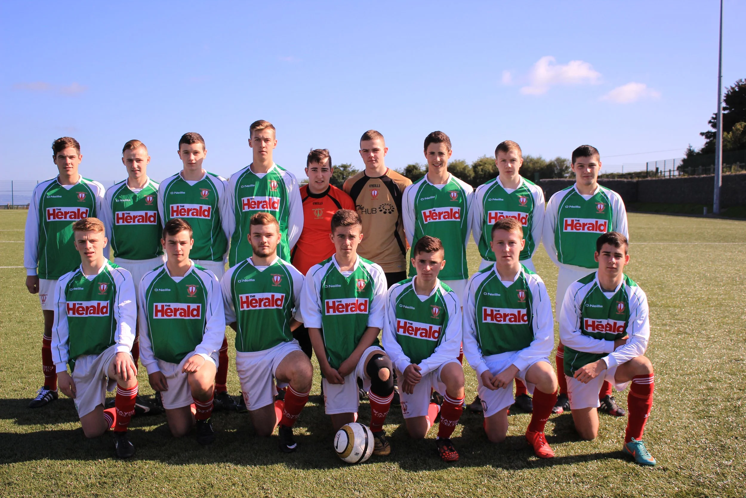 A group of young male soccer players in green and white uniforms posing for a team photo on a grassy field under a clear blue sky.