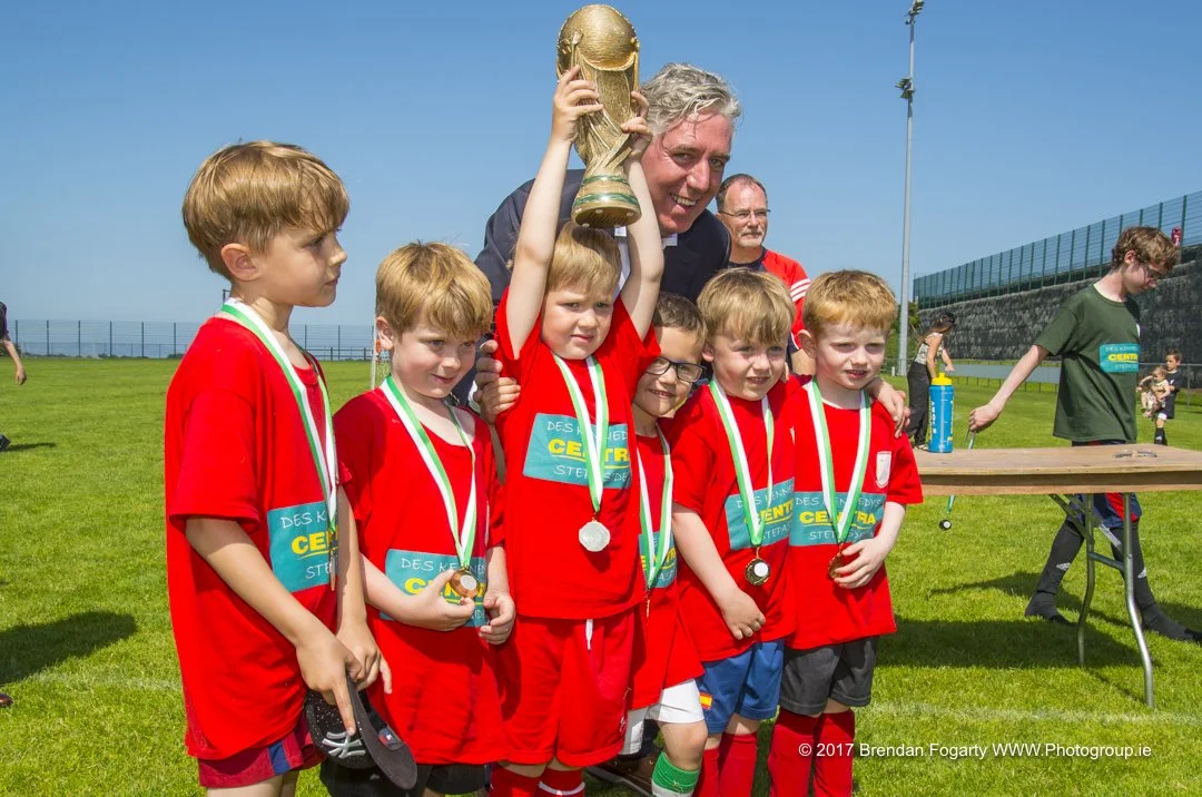 Children wearing red sports jerseys and medals, holding a trophy, standing on a sports field with an adult coach or presenter, sunny day.