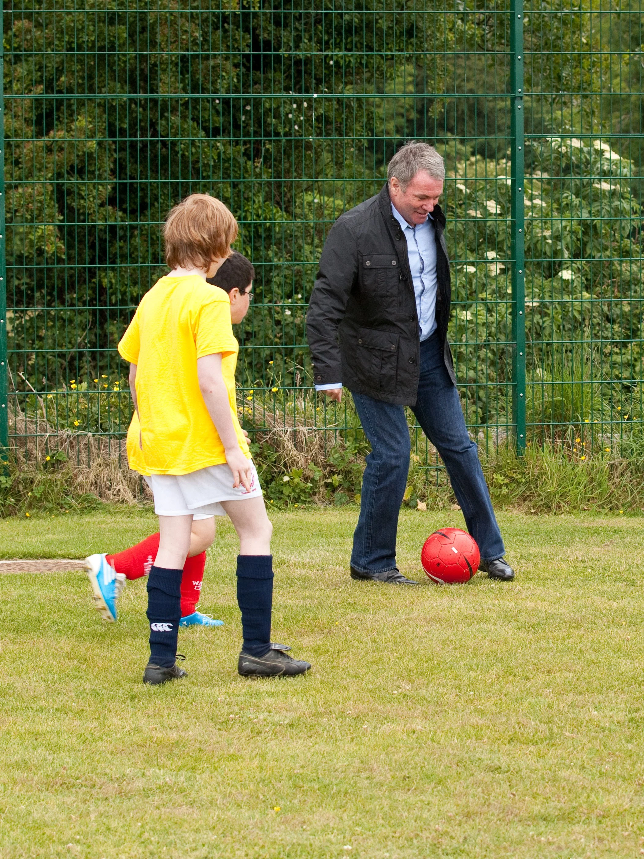 A man and two children playing soccer outdoors on a grassy field, with green trees and a metal fence in the background.