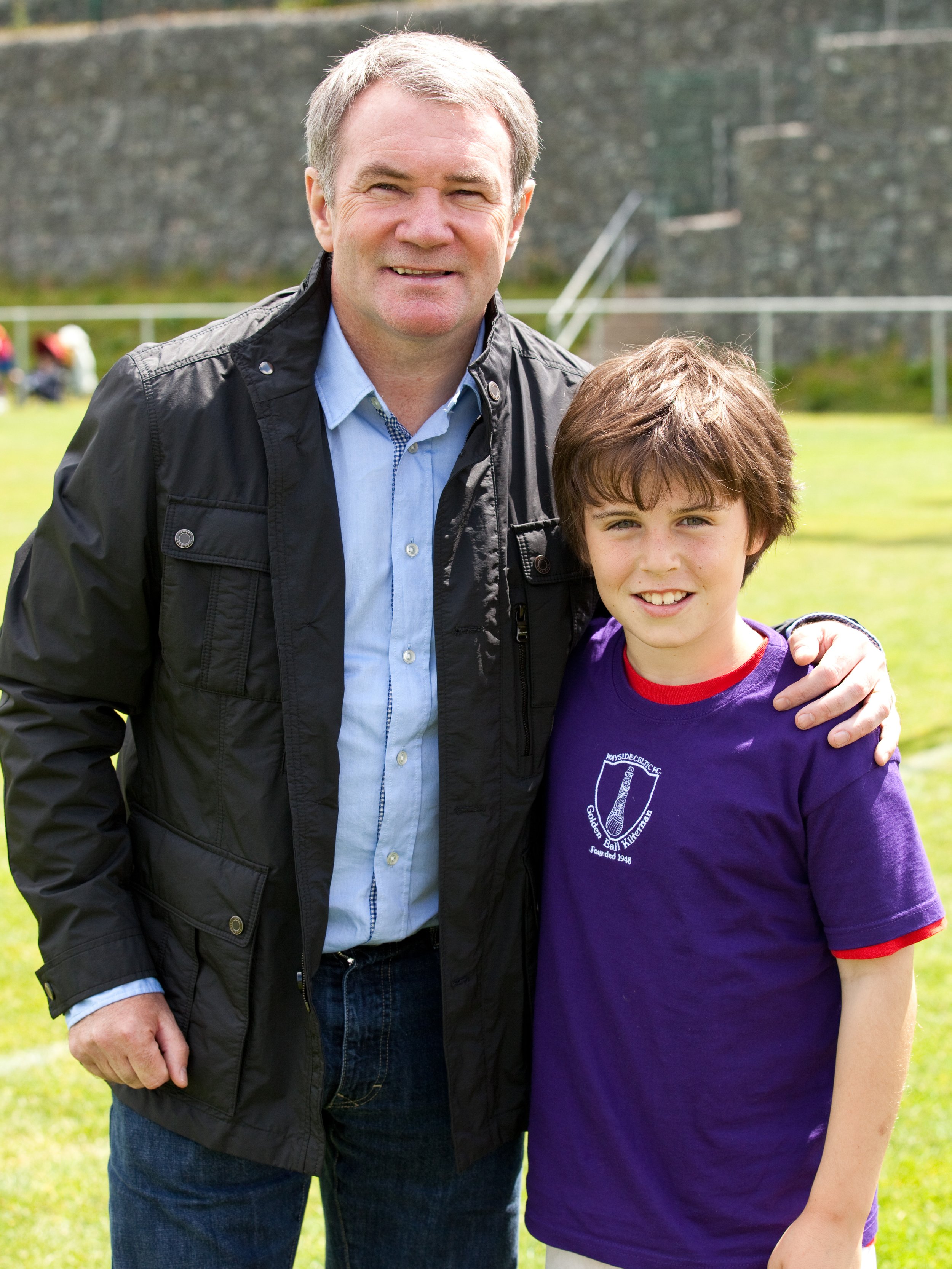 A man and boy standing outdoors, smiling. The man has grey hair and is wearing a dark jacket over a blue shirt. The boy has brown hair and is wearing a purple t-shirt with a logo on it. The background shows a grassy area and a stone wall.