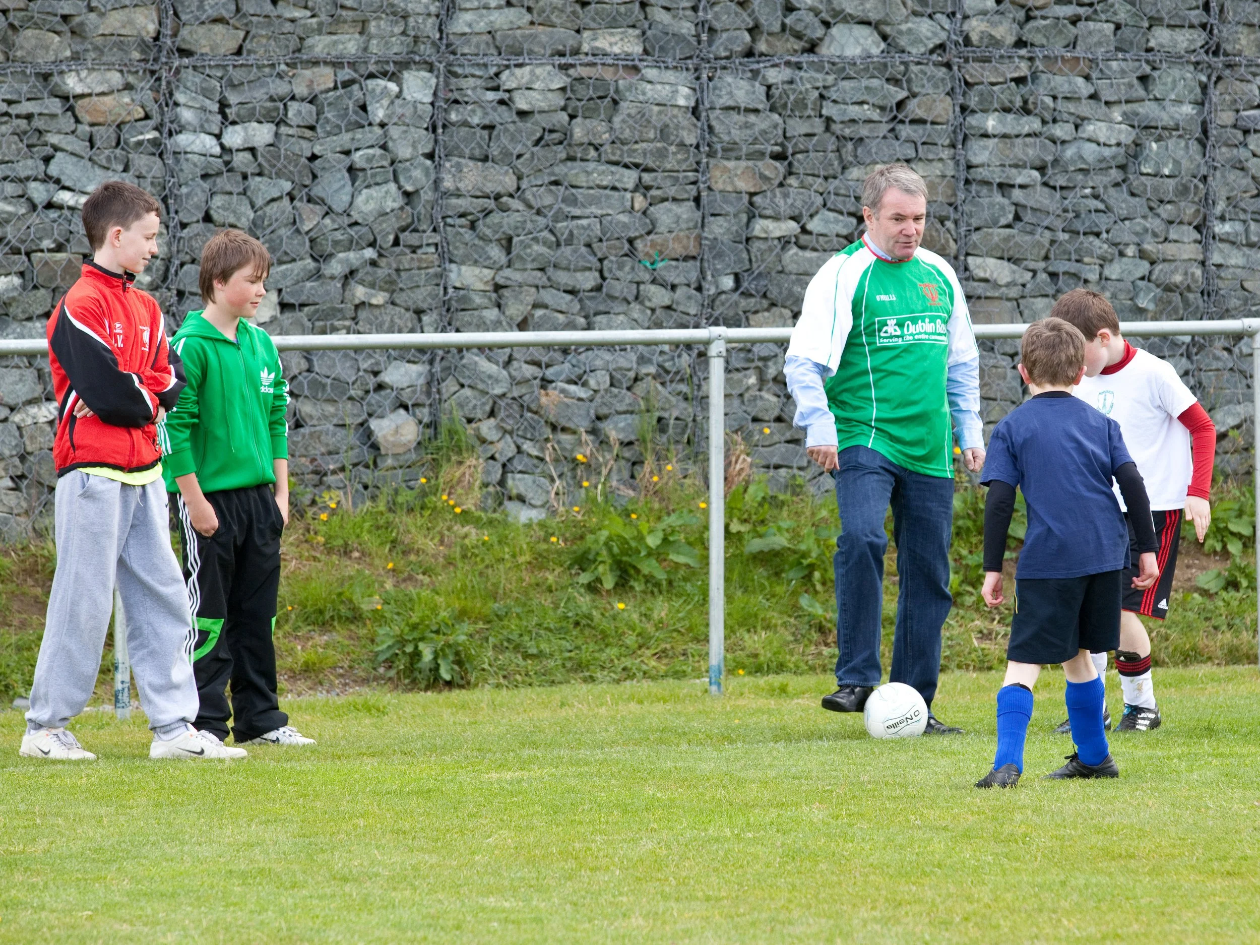 Young boys and a man standing on a soccer field near a fence, with one boy preparing to kick a soccer ball.