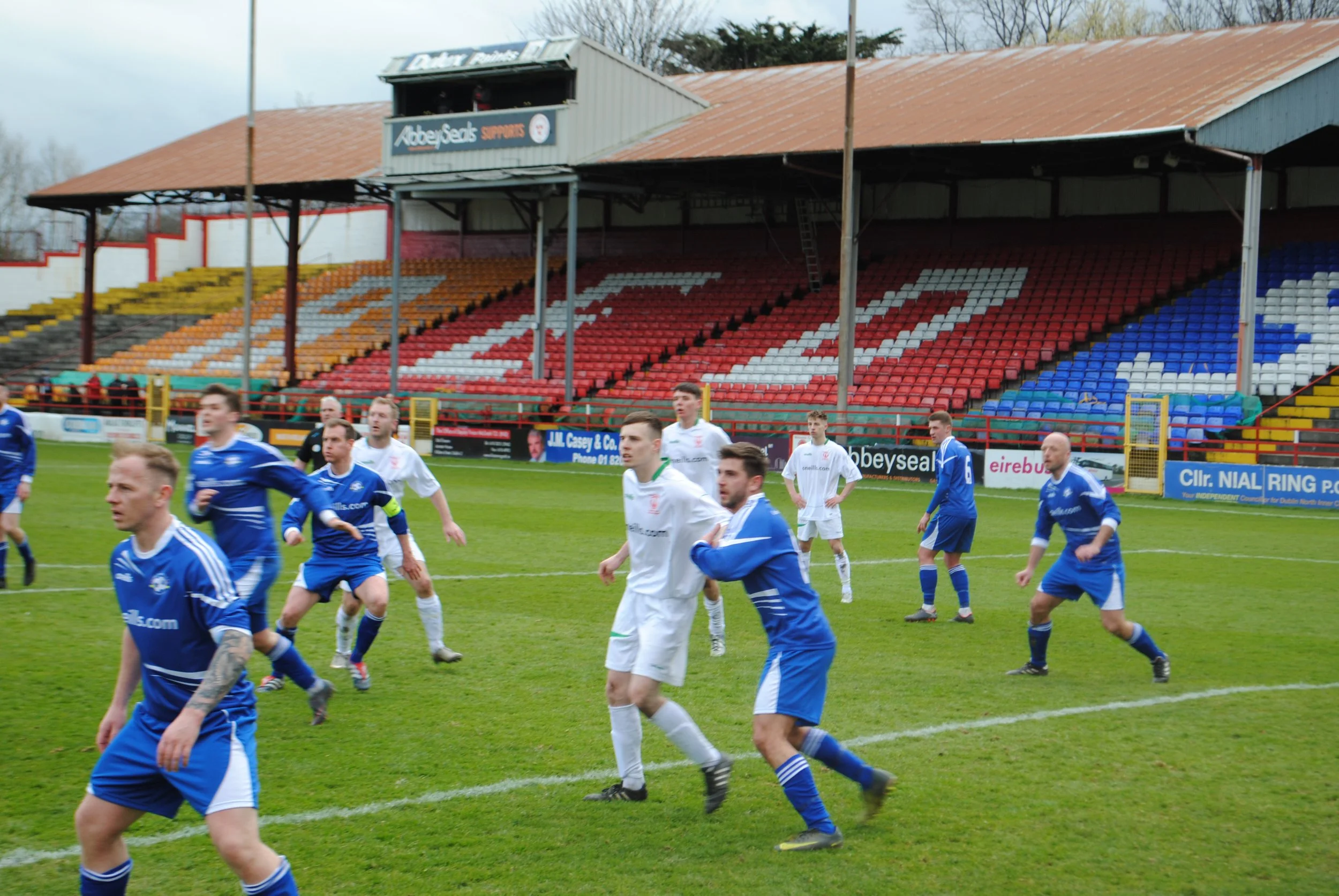 Soccer match with players in blue and white uniforms on a field, with an empty grandstand with red, white, and blue seats in the background.