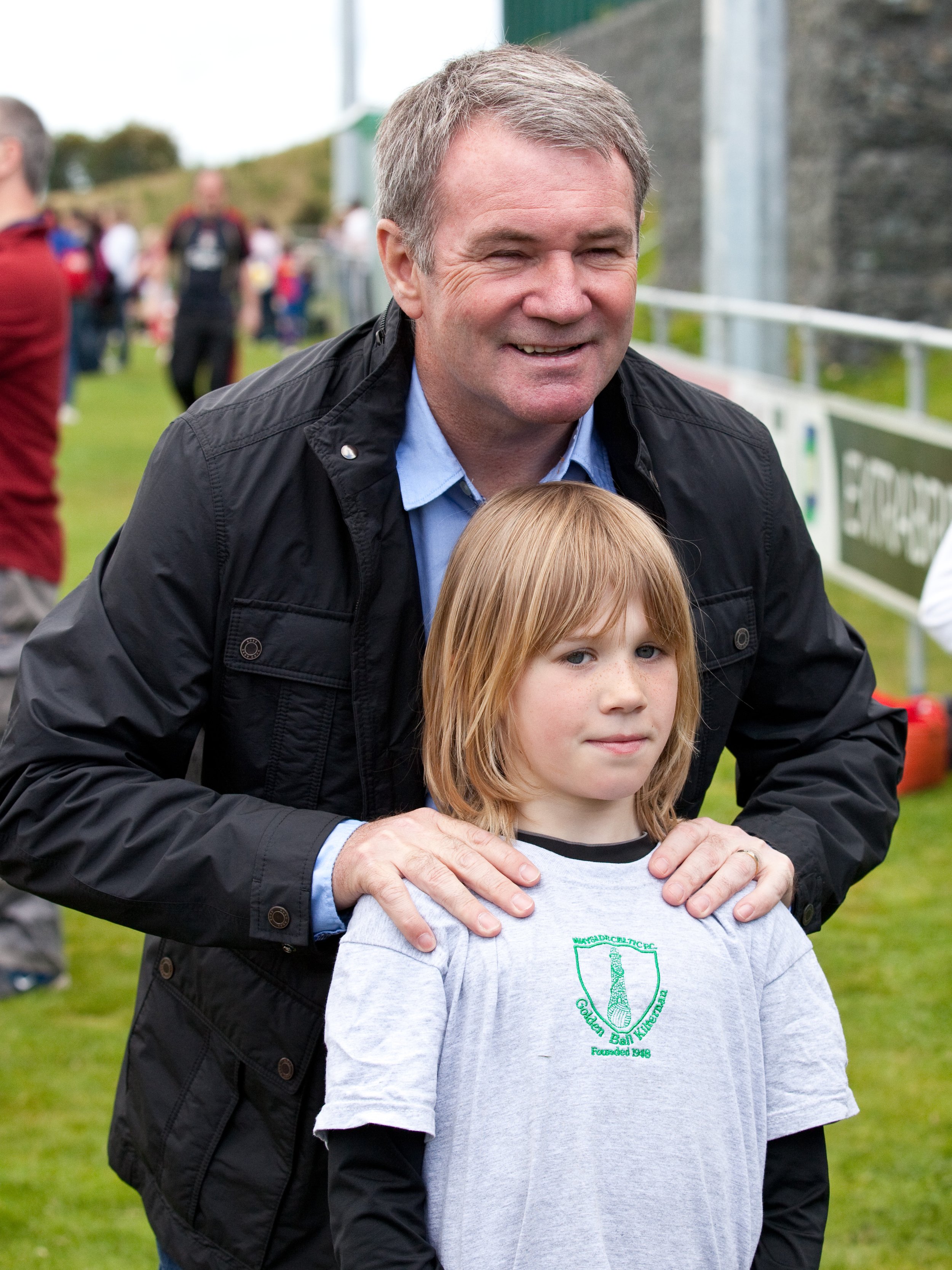 A smiling man with short gray hair and a black jacket standing behind a young girl with red hair and blue eyes, outdoors at a sports event with other people in the background.