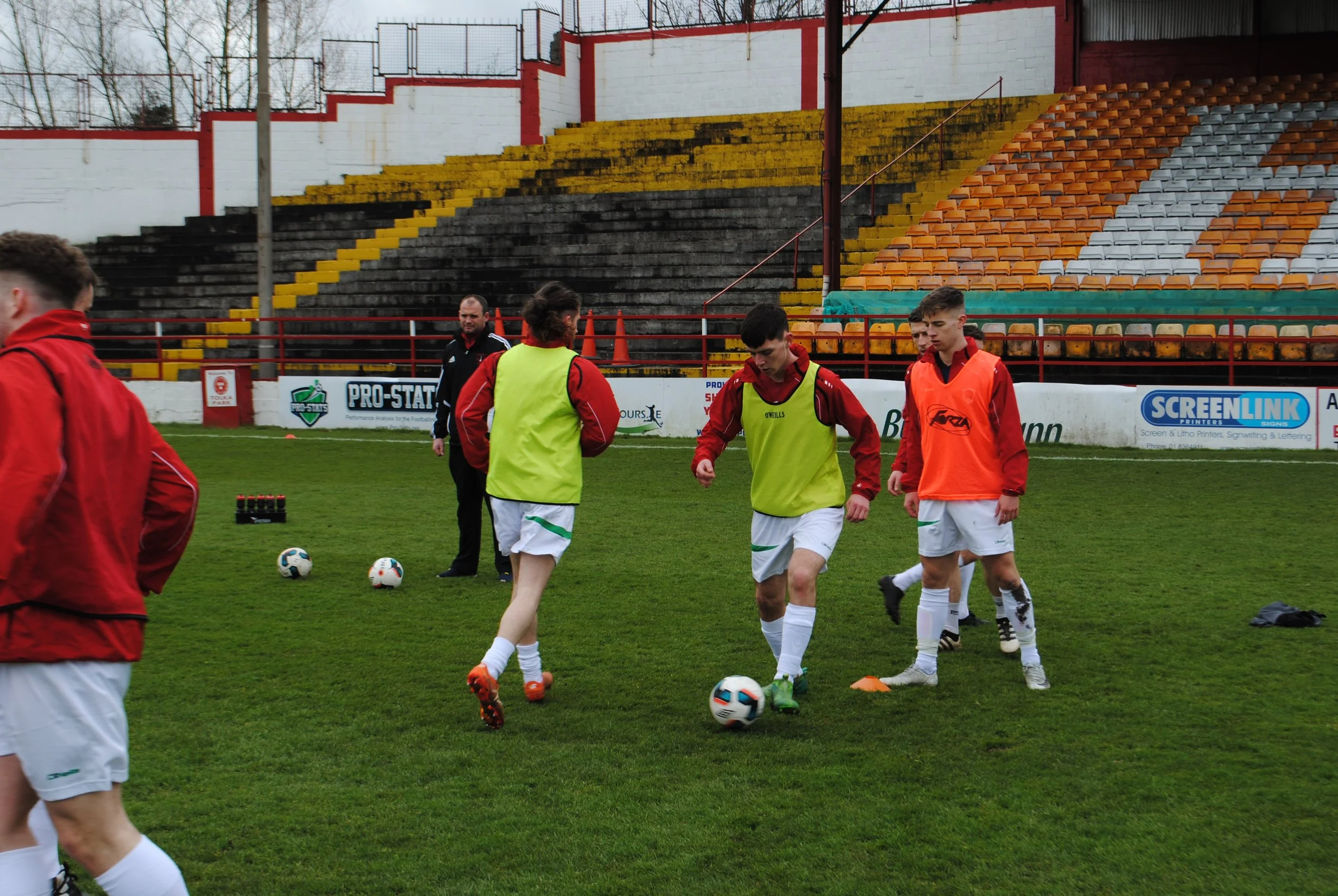 Soccer players practicing on a field, some wearing green and orange training vests, with a coach in black, in a stadium with yellow, black, white, and orange seats and advertisements along the sidelines.
