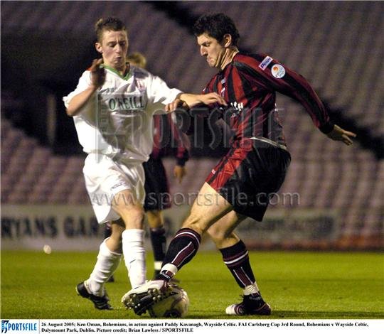 Two soccer players compete for the ball on the field during a match, one in a white uniform and the other in a black and red uniform, with empty stadium seats in the background.