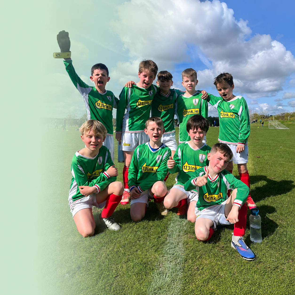 Group of young boys in green and white soccer uniforms posing on a grassy field with cloudy sky in the background, some crouching and some standing, celebrating after a game.