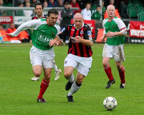 Four soccer players racing towards a ball on a soccer field, with the background of spectators.