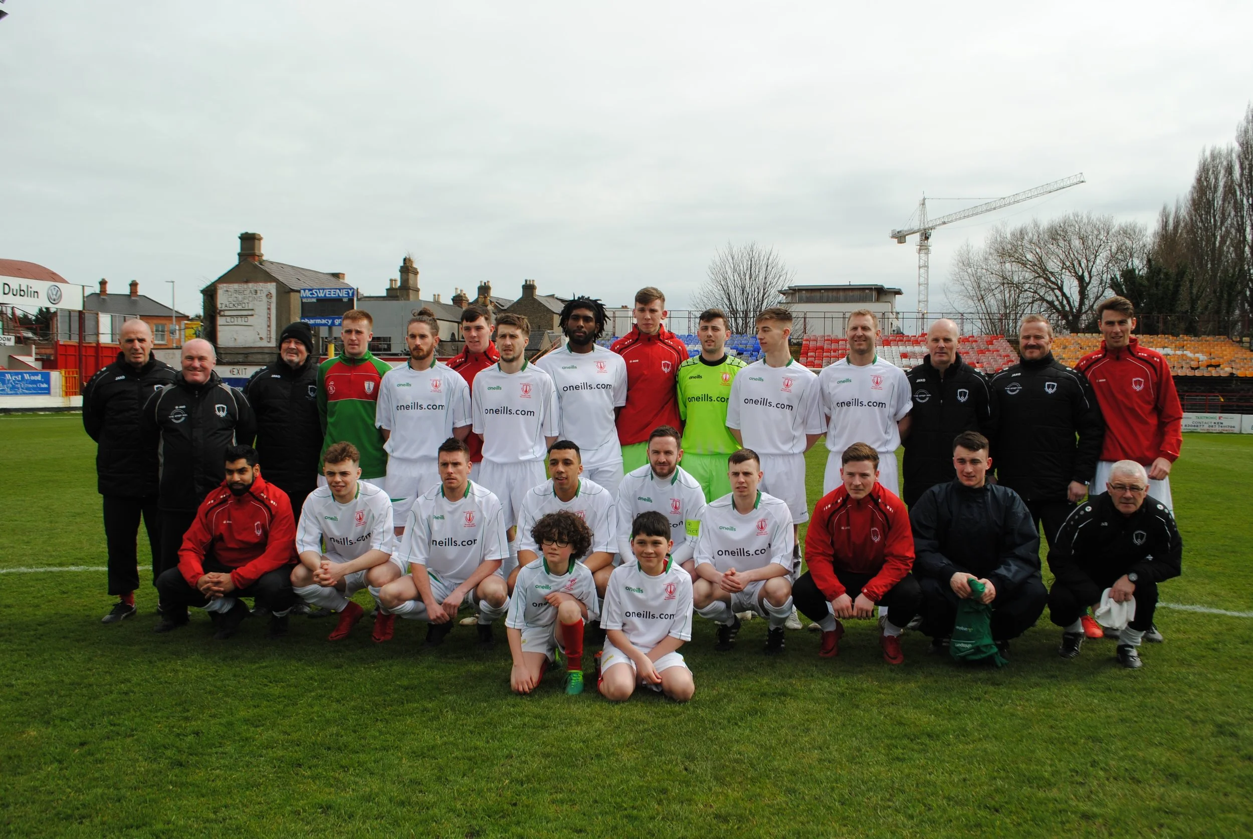 A group of soccer players and coaches posing for a team photo on a soccer field with stands and trees in the background.