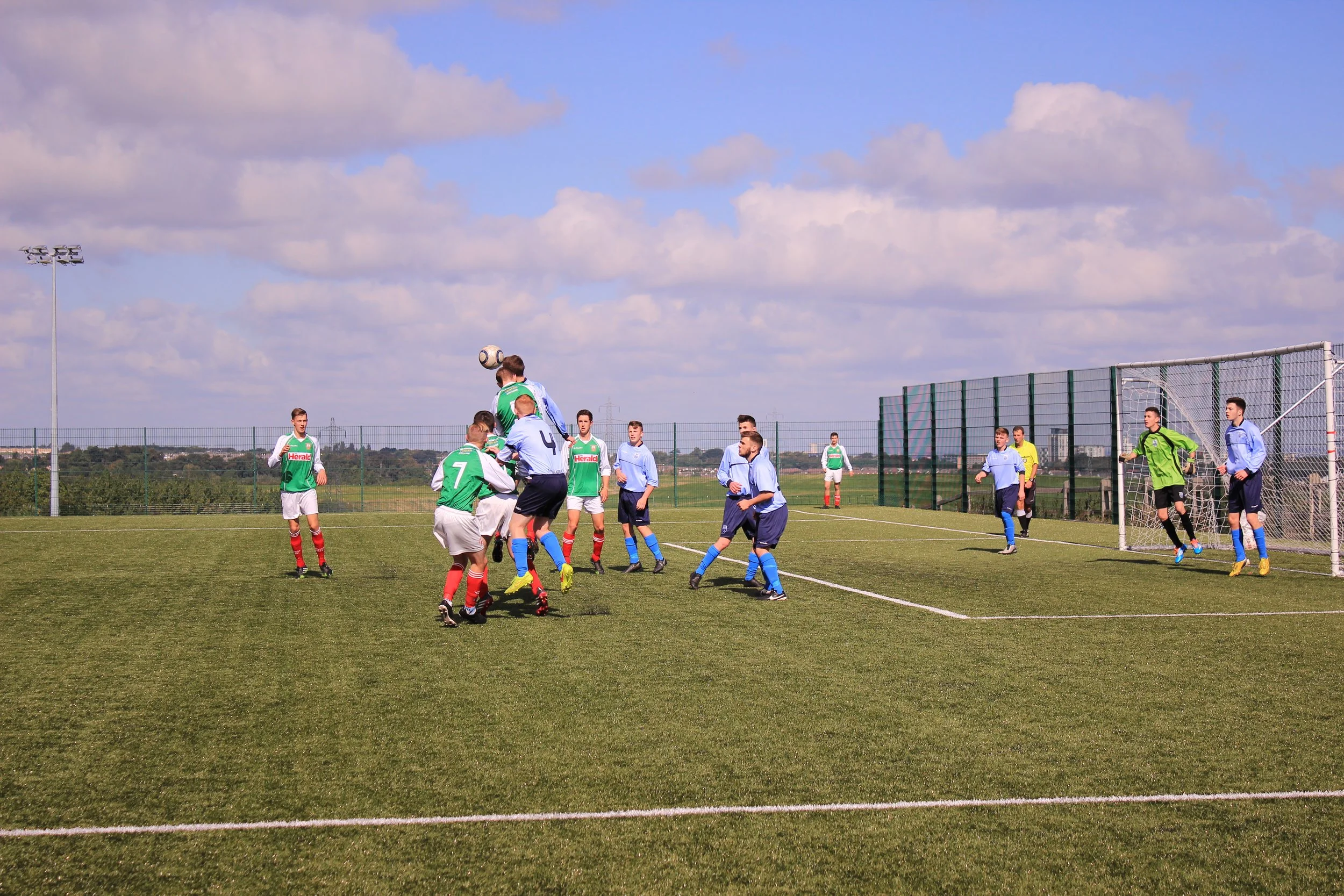 A group of young soccer players on a green field during a match, with some players jumping for the ball near the goal, and others standing and watching under a partly cloudy sky.