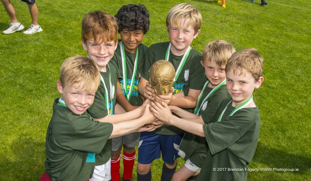 Children in green sports jerseys holding up a gold soccer trophy on a grass field.