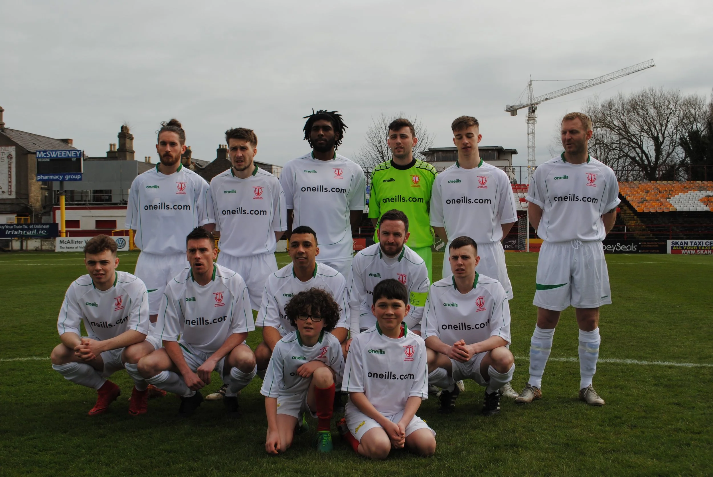 Group of young male soccer players in white uniforms posing on a soccer field, with a few young children kneeling in front, under an overcast sky with some buildings and a construction crane in the background.