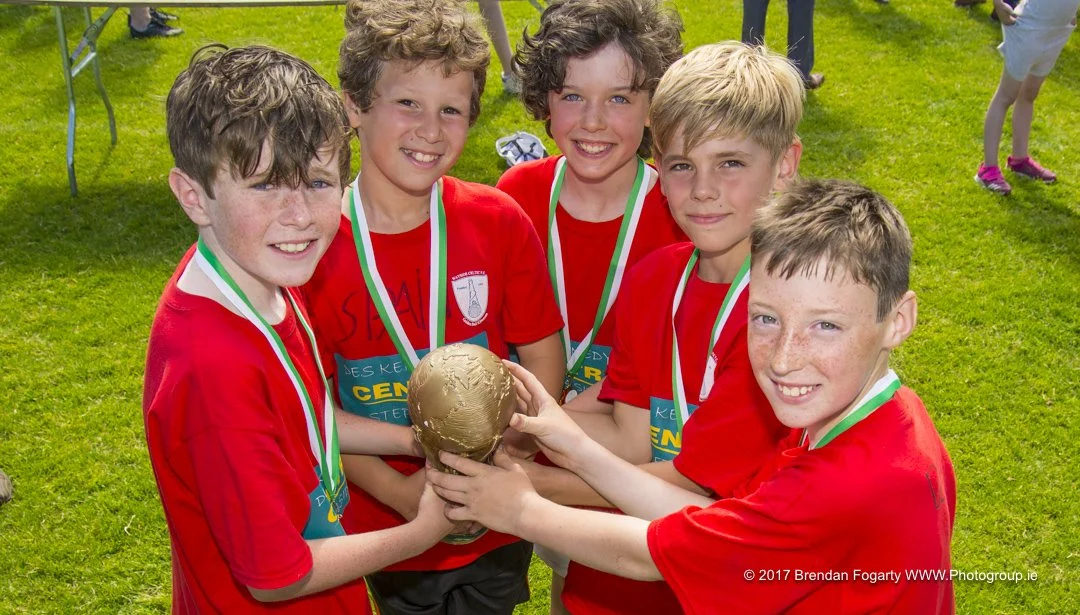 Group of five young boys in red sports uniforms holding a gold-colored trophy and medal in a grassy outdoor setting.
