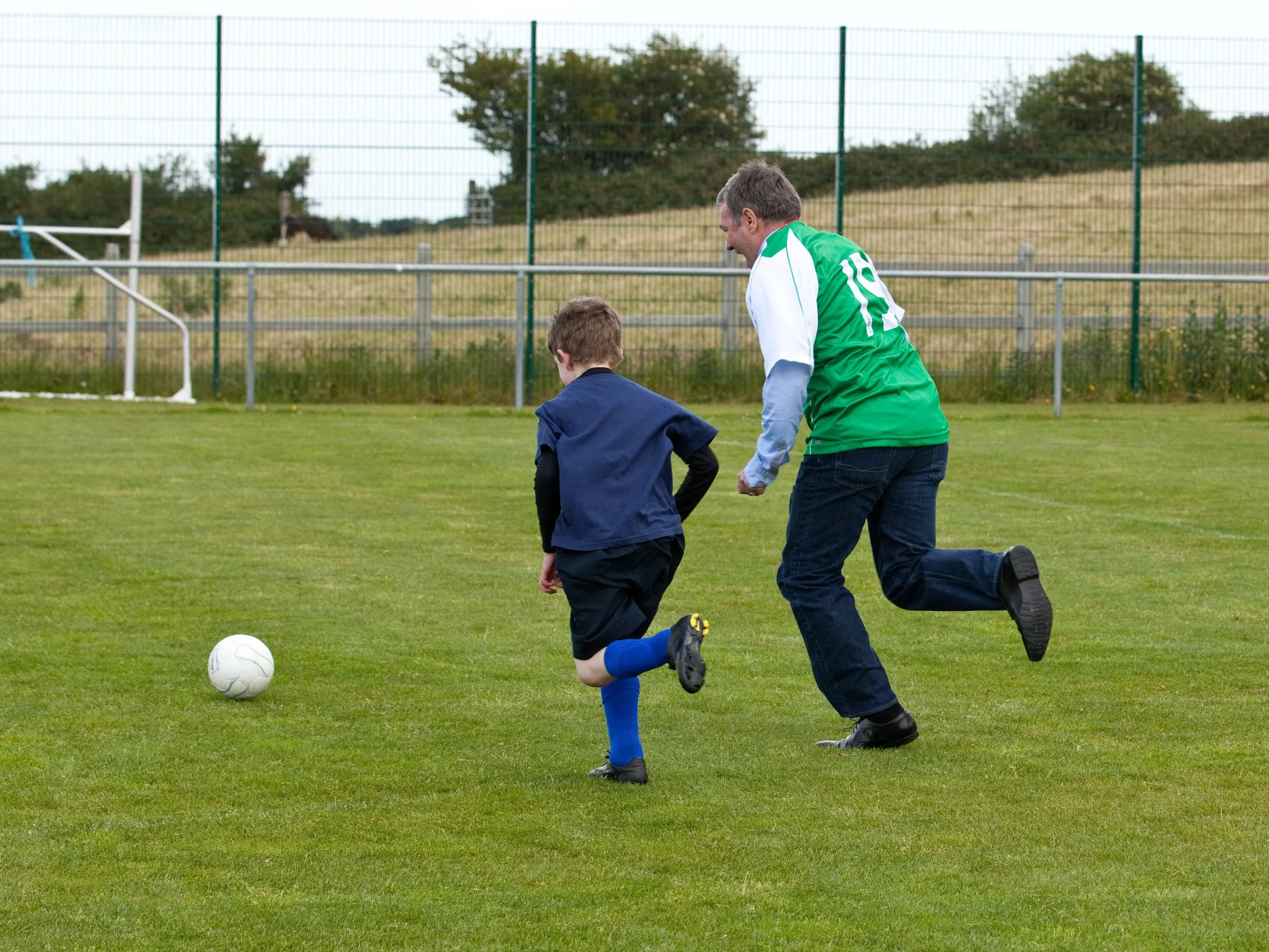 A man and a young boy are playing soccer on a grassy field. The man, wearing a green and white shirt with the number 10, is running alongside the boy, who is in a blue uniform. They are both focused on the white soccer ball on the ground, with trees,