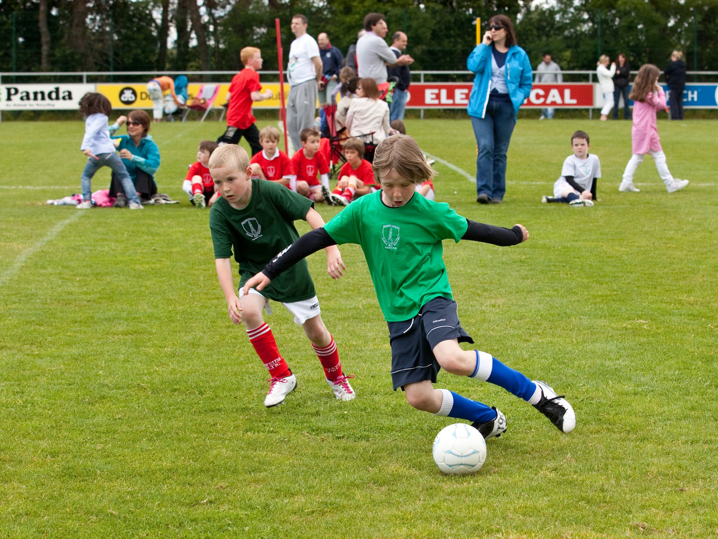 Two young boys compete for a soccer ball on a grassy field during a youth soccer game, with spectators and players in the background.