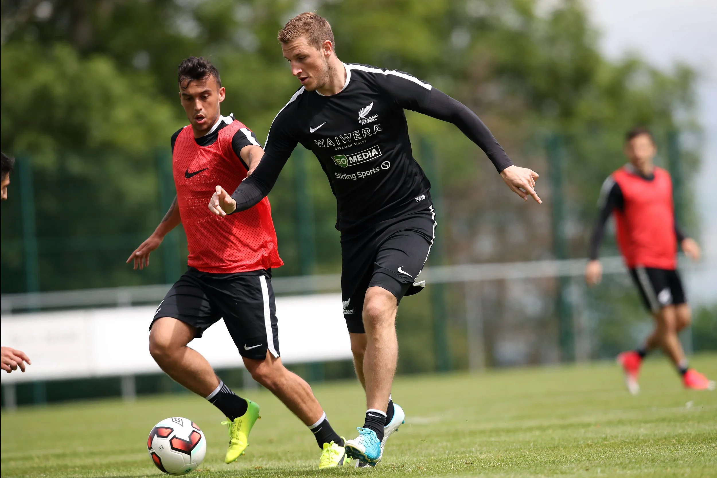 Two soccer players competing for the ball on a field. One player in black jersey and black shorts, the other in red jersey and black shorts. The black jersey has white text and a logo that reads 'WAIWERA' and 'NZ FOOTBALL'. The scene is outdoors with