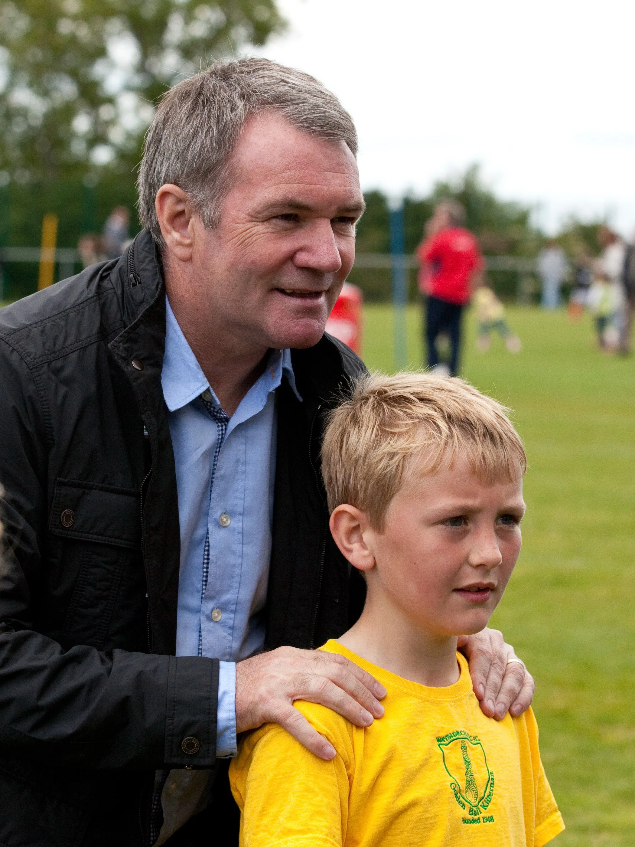 An adult man with gray hair, wearing a black jacket and blue shirt, is standing outdoors with a young boy with red hair, wearing a yellow sports shirt. The man has his arm around the boy, and both are smiling. In the background, there are people on a