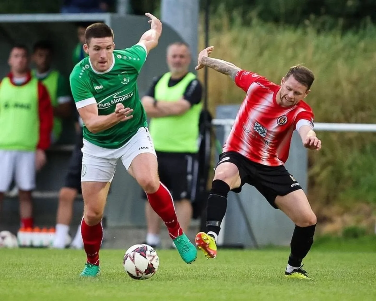 Two soccer players competing for the ball during a match, one in a green jersey and the other in a red and white jersey, with players and spectators in the background.