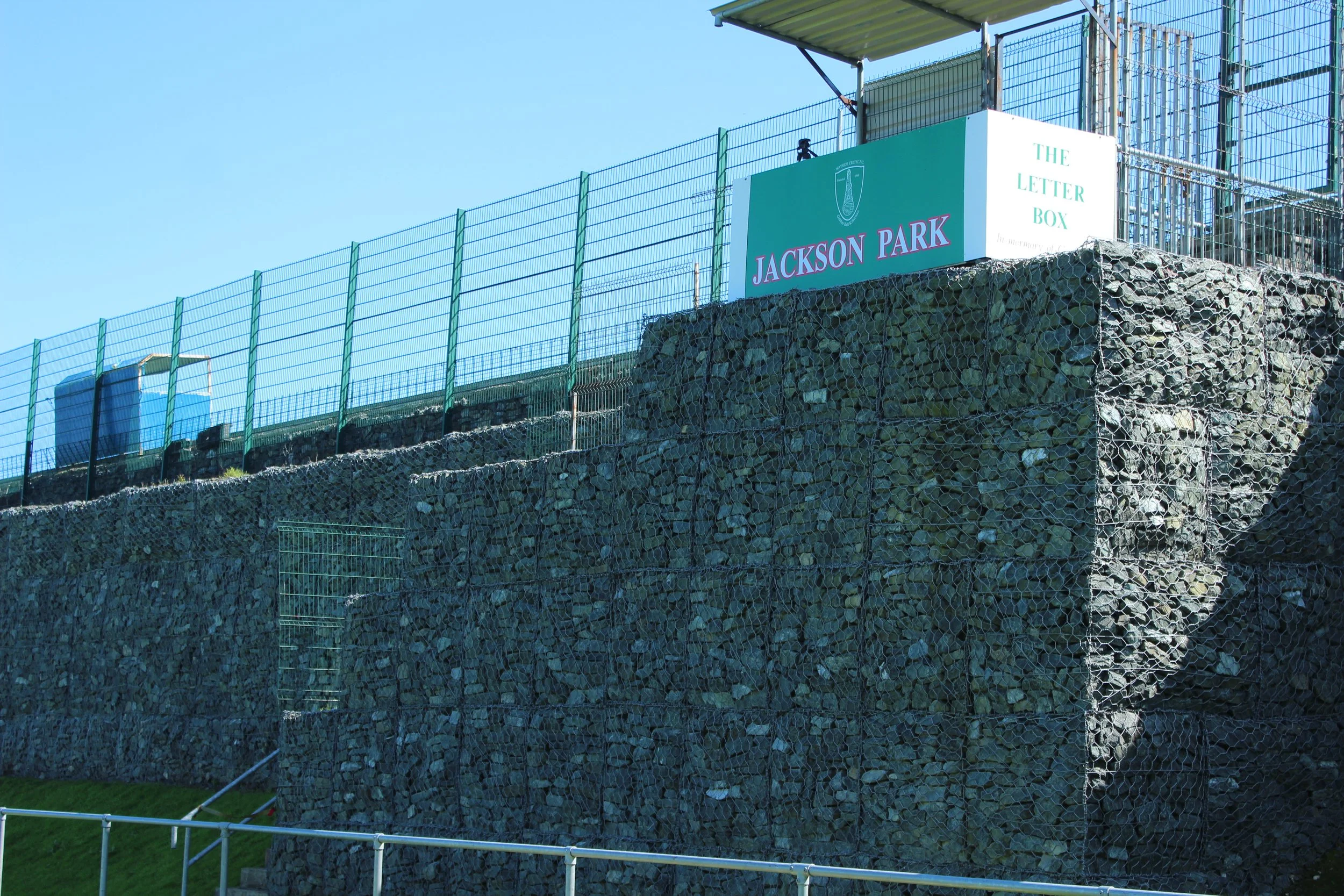 A portion of a sports stadium or park with a tall green fence, a sign reading "Jackson Park" and "The Letter Box," and a stone retaining wall, under a clear blue sky.
