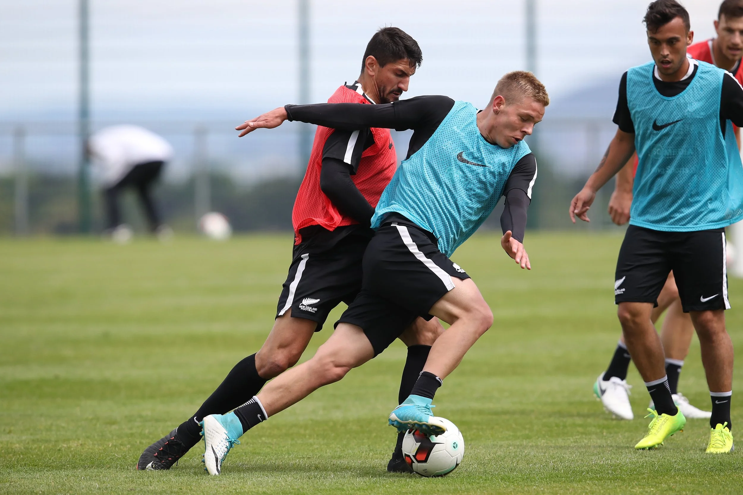 A group of soccer players practicing on a green field, with one player in a blue vest controlling the ball while others watch and prepare to play.