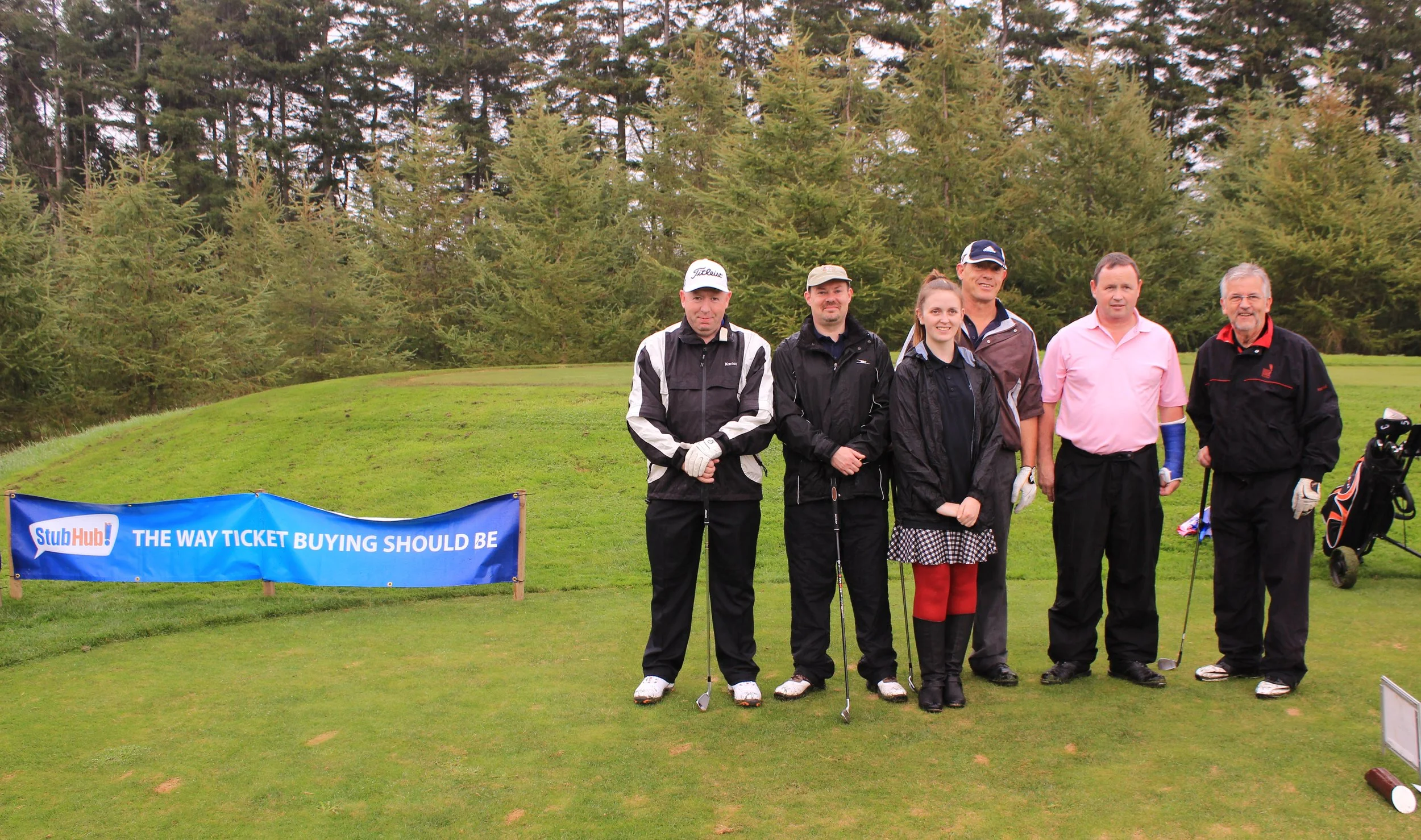 Group of five people standing on a golf course, smiling, with trees in the background. There is a blue sign that reads, 'StubHub! The way ticket buying should be'.