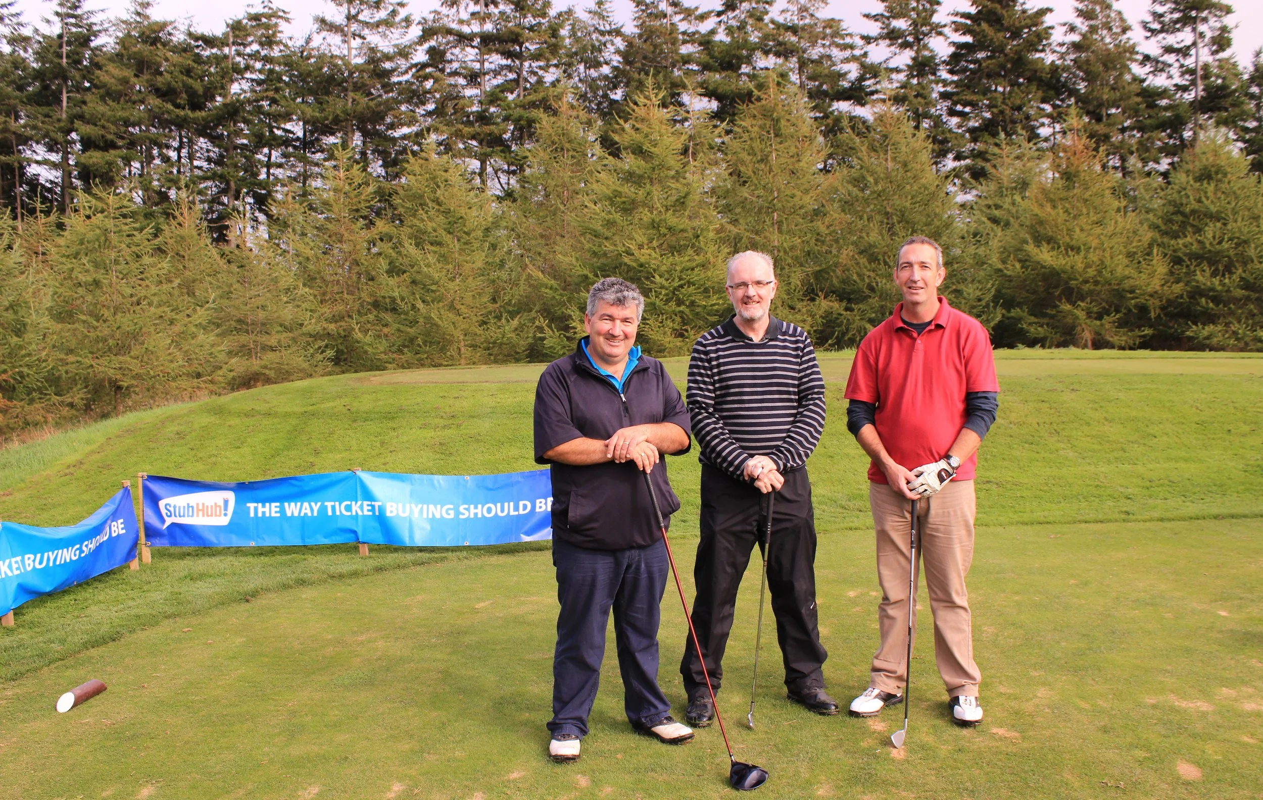 Three men standing on a golf course holding golf clubs. They are smiling and dressed in golf attire. A banner in front of them reads, 'StubHub! THE WAY TICKET BUYING SHOULD BE.' In the background, there are trees and a grassy hill.