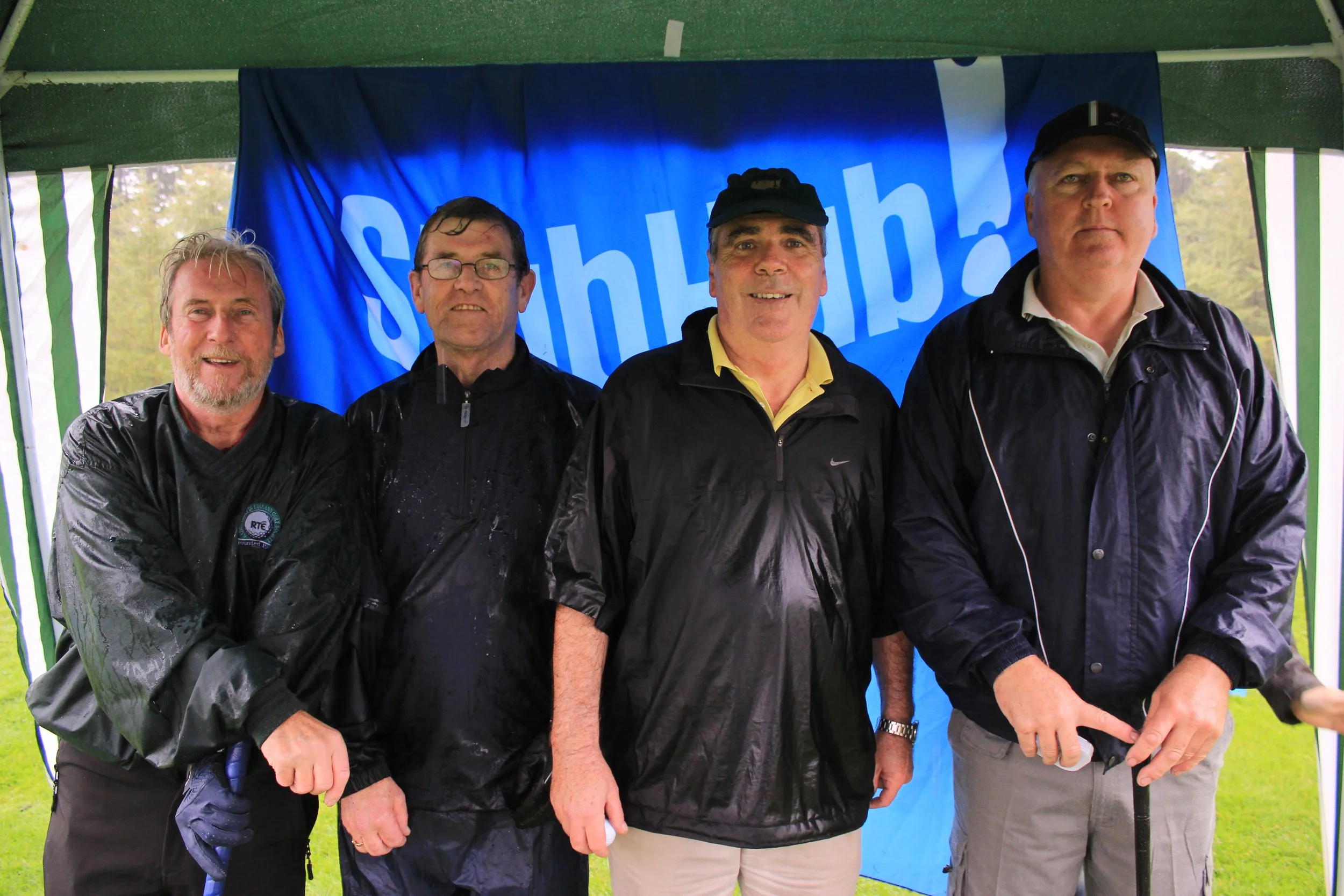 Four men standing outdoors in front of a blue banner with white text, wearing black rain jackets and hats, across a green and white striped tent.