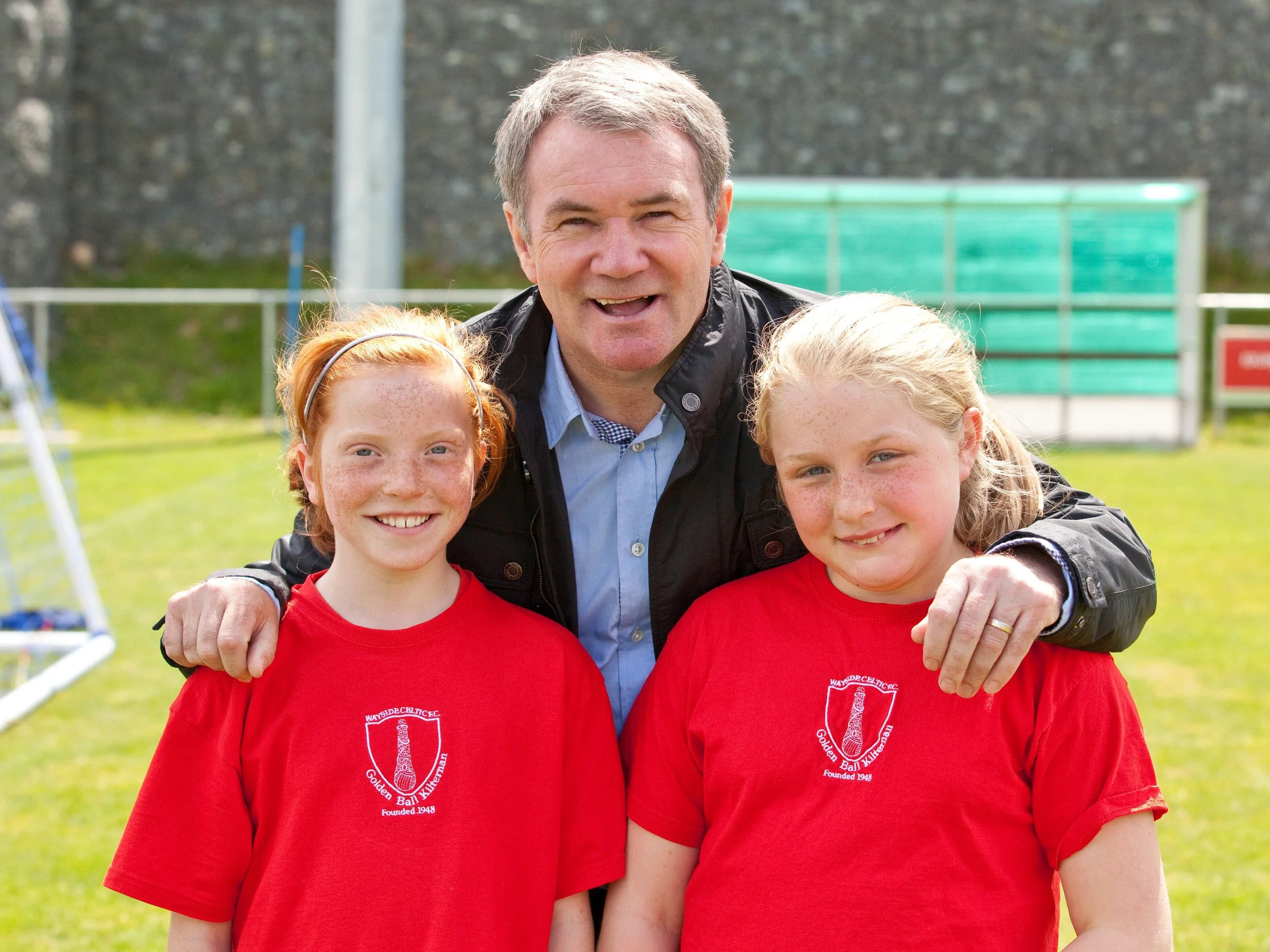 A man with short gray hair and wearing a black jacket is posing outdoors with two young girls with red hair and freckles, all smiling at the camera. The girls are wearing red shirts with a white emblem on the front that reads 'Wayside Bible Christian