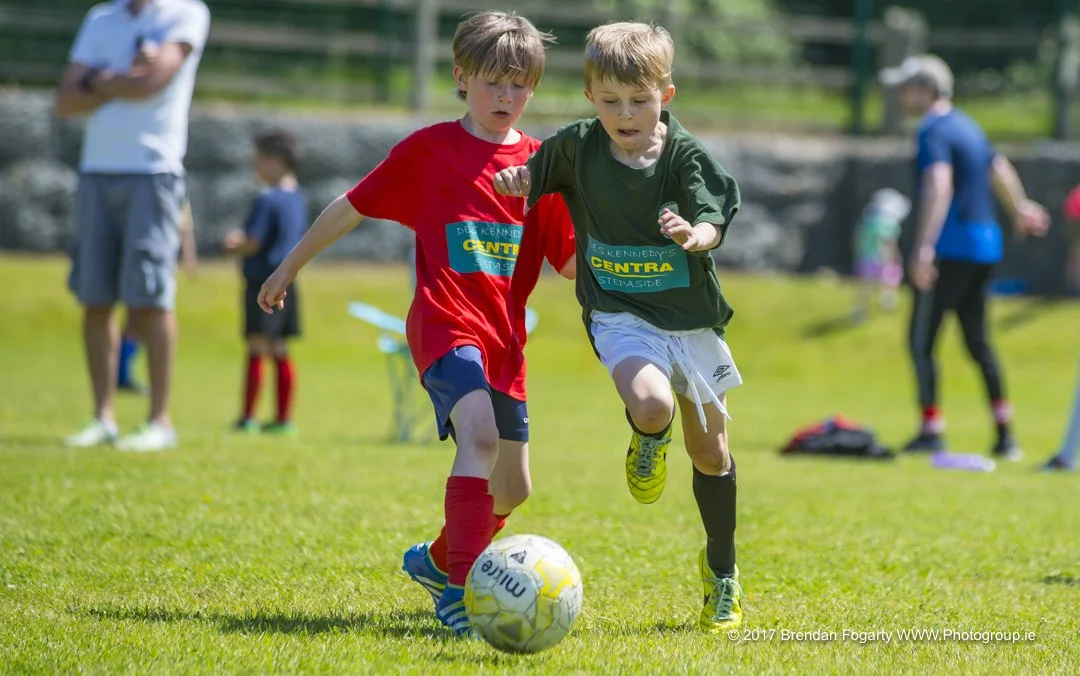 Two young boys playing soccer outdoors on a sunny day, chasing a ball with other children and adults in the background.