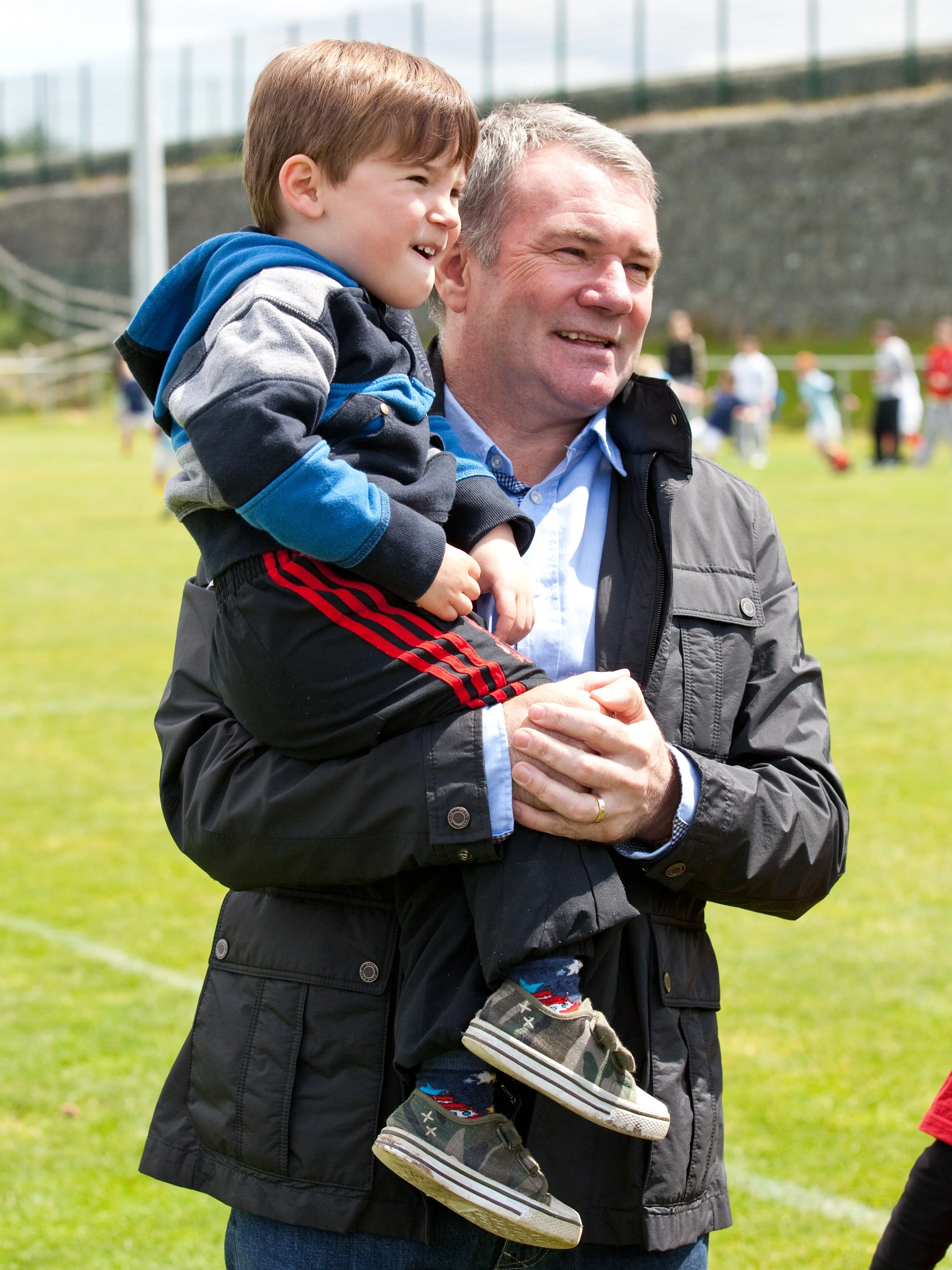 A man holding a young boy on his shoulder outdoors at a park or sports field on a sunny day.