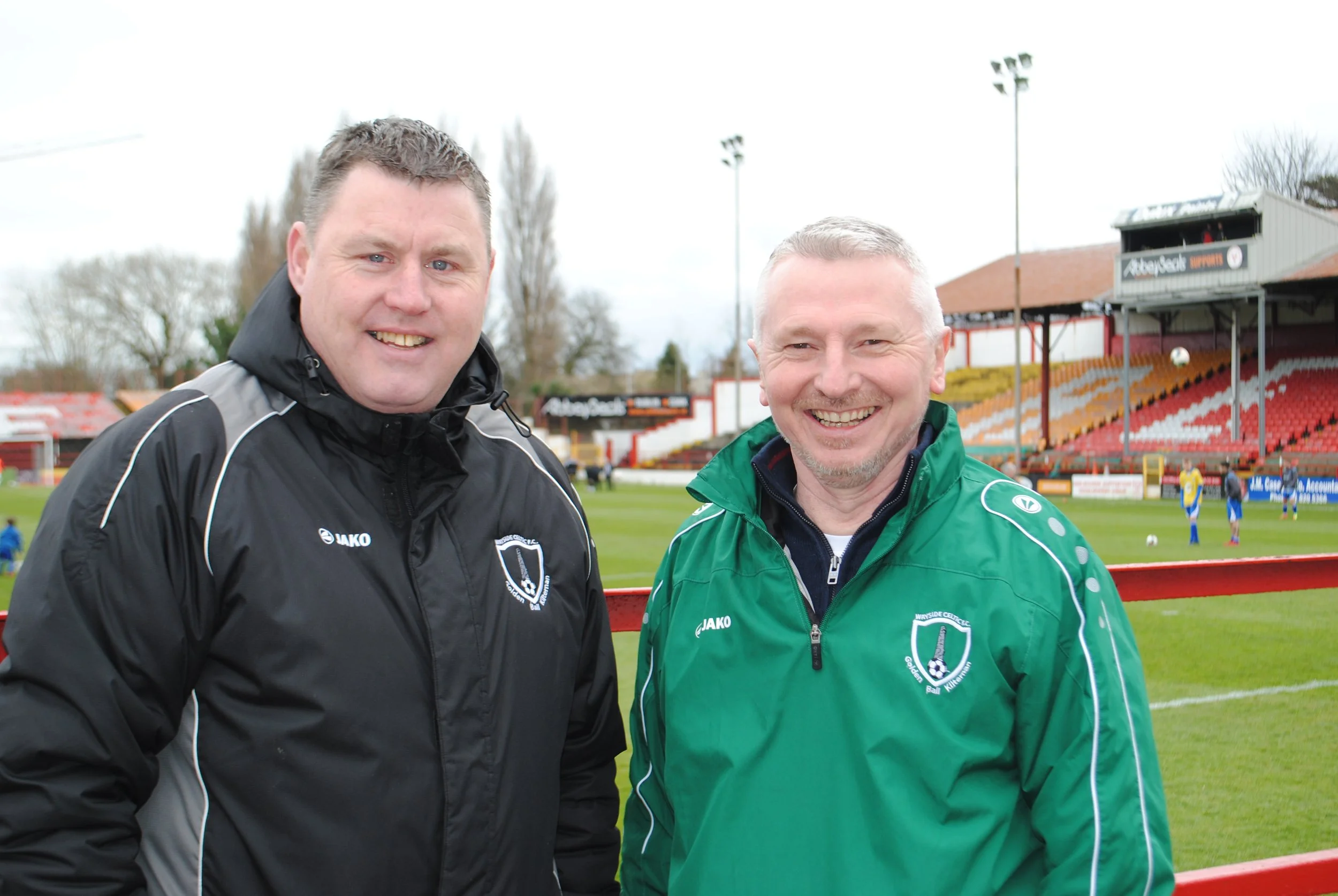 Two smiling men in sports jackets standing on a football field with a stadium and players in the background.