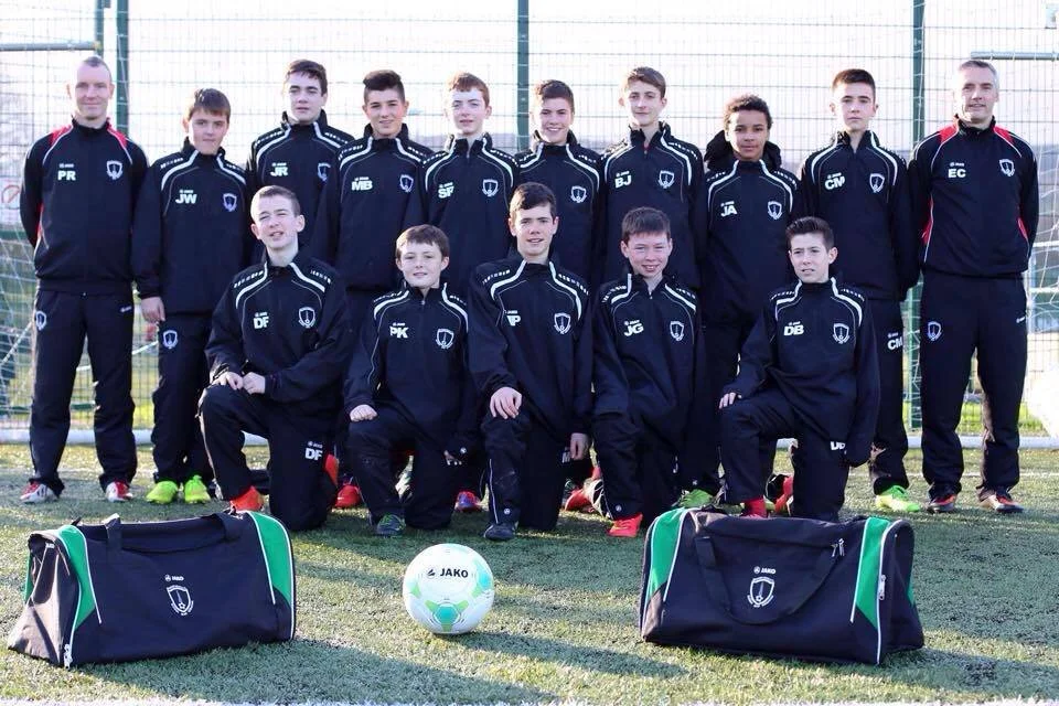 Youth soccer team and coaches posing for a team photo on a field, with soccer bags and ball in front, and a soccer goal in the background.