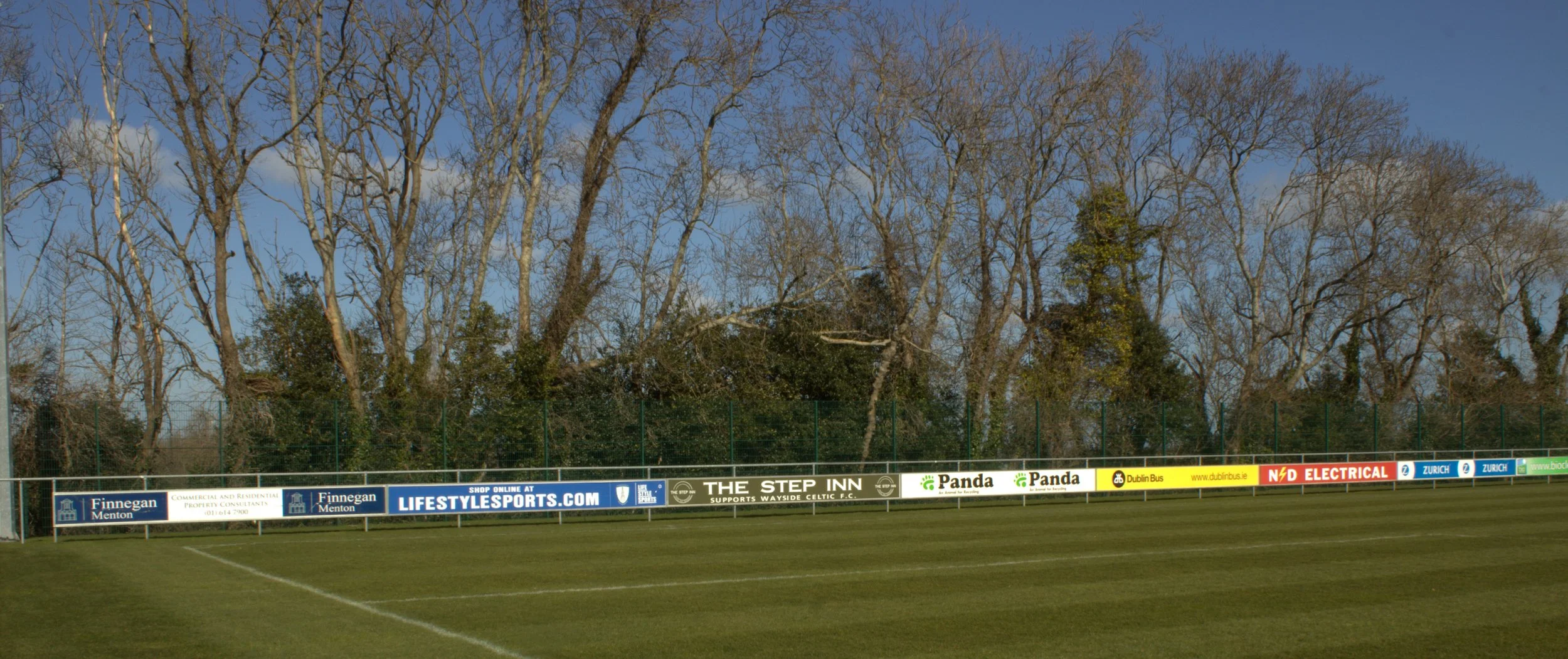 A view of a soccer field with a green grass playing surface, advertising banners along the fence, and tall, leafless trees in the background under a clear blue sky.