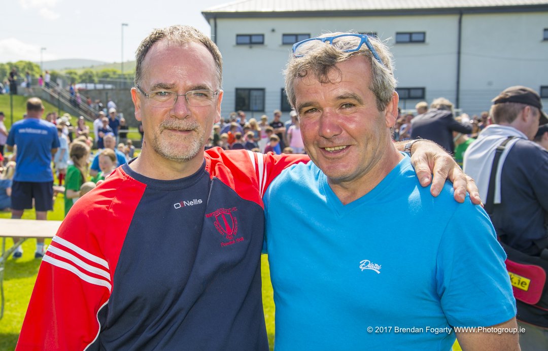 Two men standing outdoors at a crowded event, smiling and embracing, one wearing glasses and a red and black sports jersey, the other wearing a blue t-shirt.