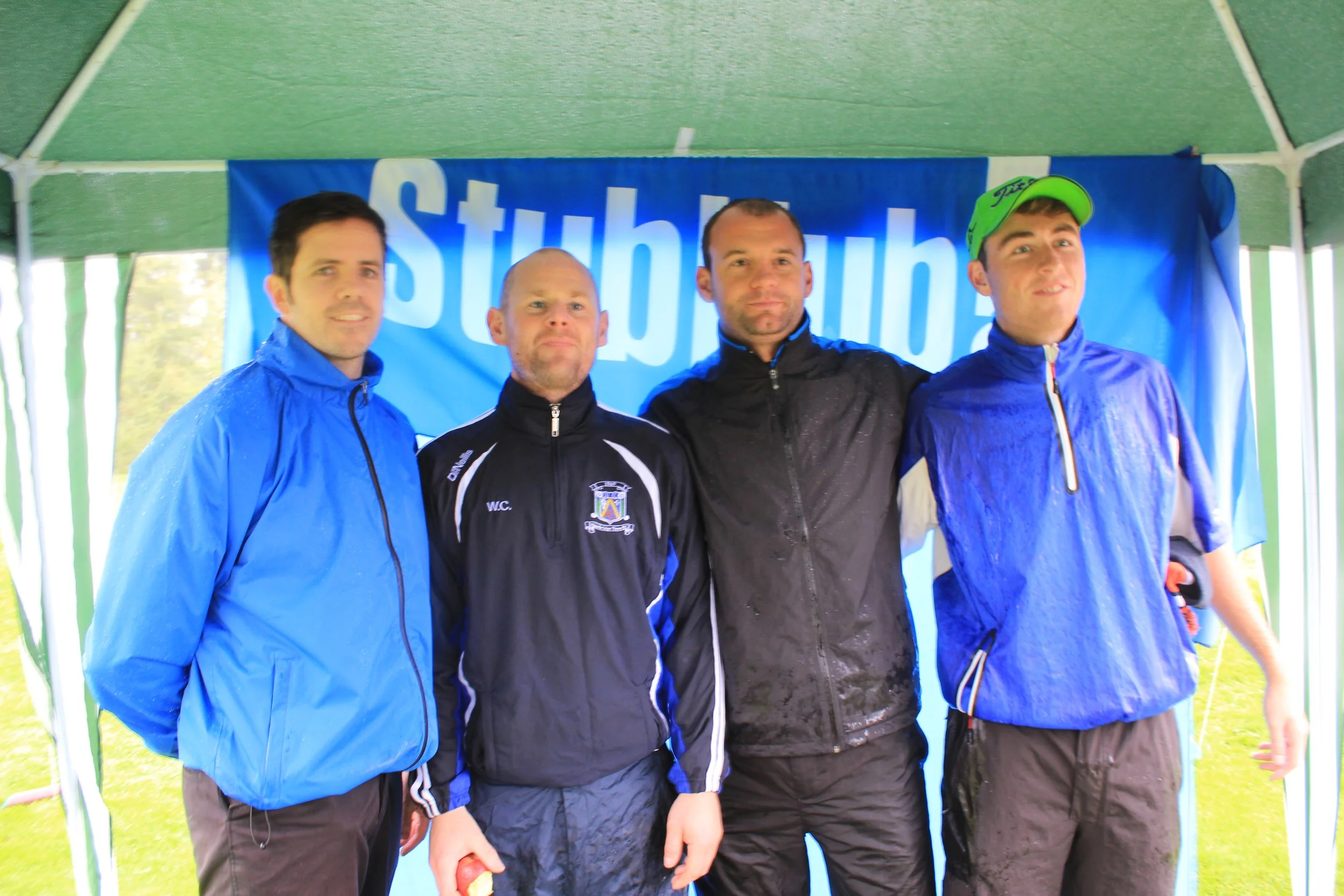 Four men standing together under a green tent, posing for a photo at an outdoor event. They are wearing sports jackets and caps, with a blue banner in the background.