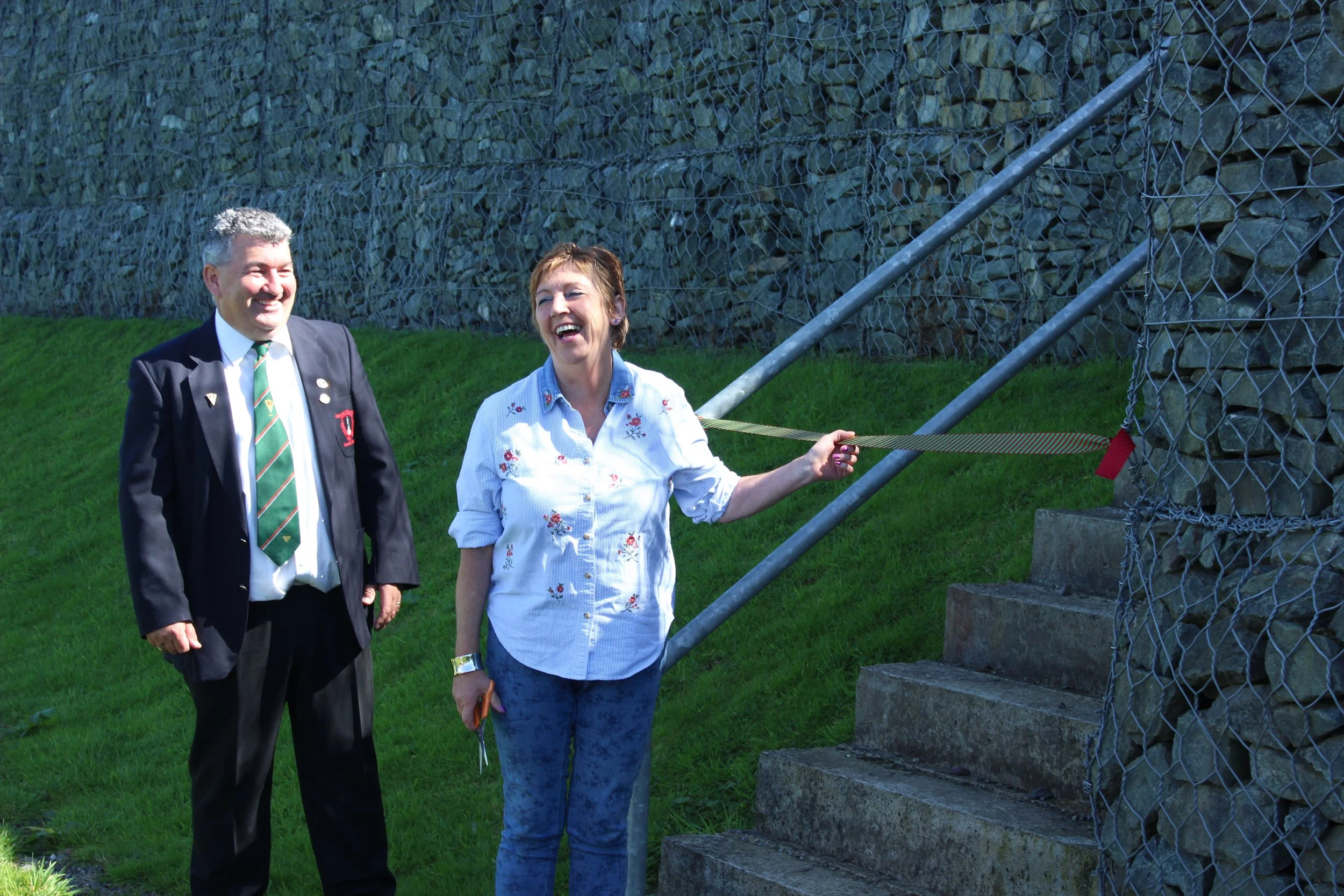 A man in a dark suit and tie, and a woman in a light embroidered blouse, are standing outdoors by a stone staircase and chain-link fence, laughing and smiling.