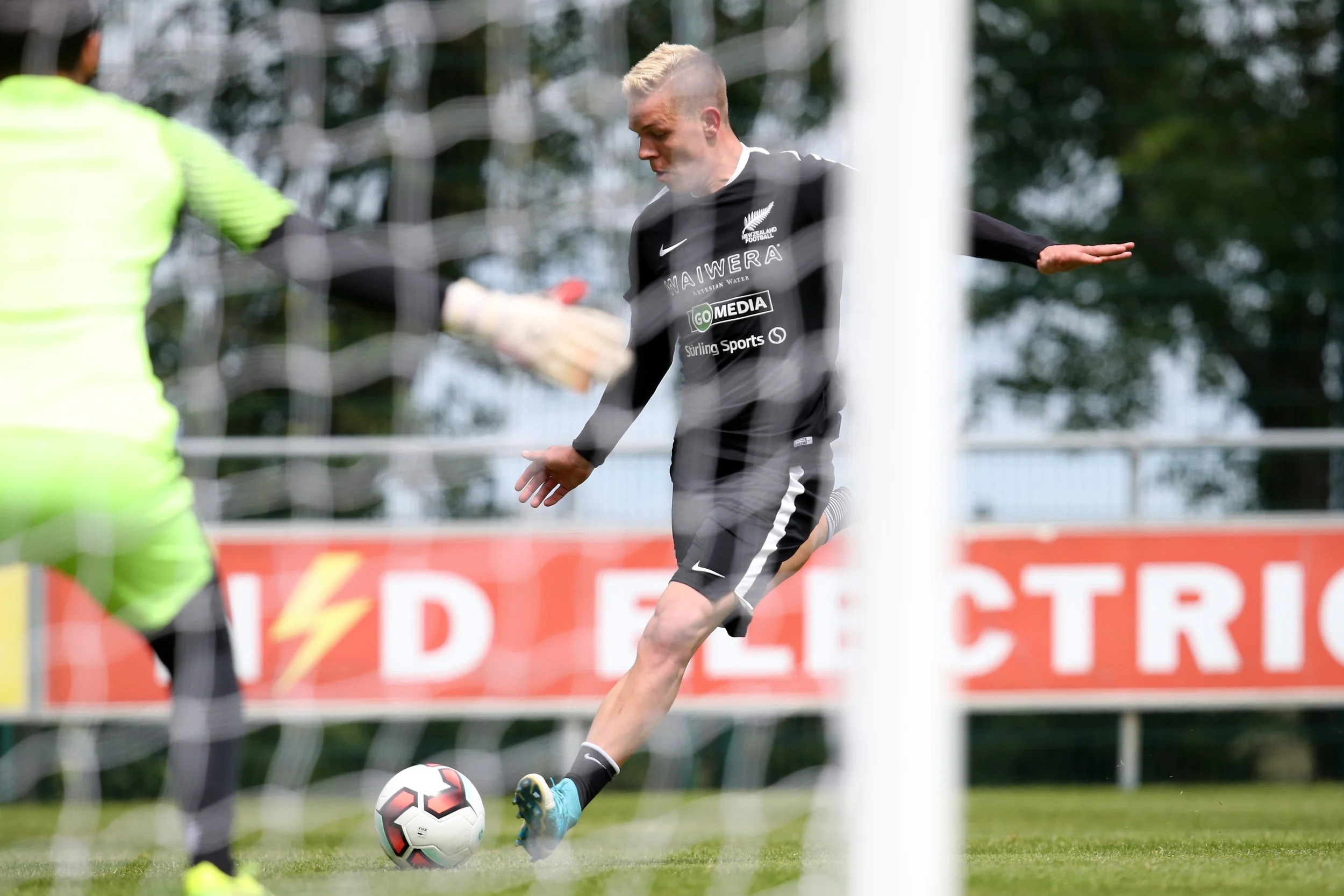 A soccer player in black uniform kicking a ball near the goal, with a goalkeeper in a lime green jersey in front, seen through the goal net.