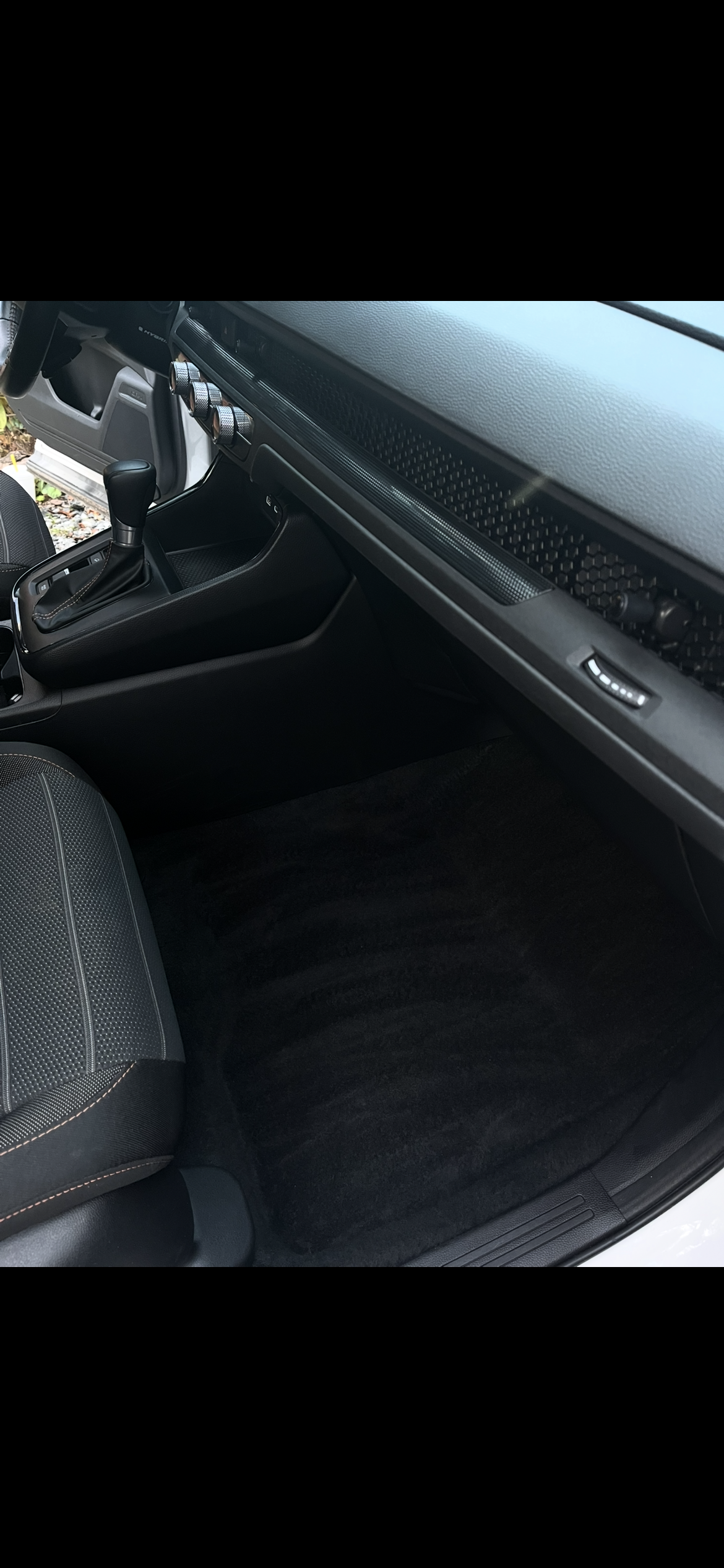 Interior of a car showing an automatic gear shift, black dashboard with four control knobs, and a black carpeted floor.