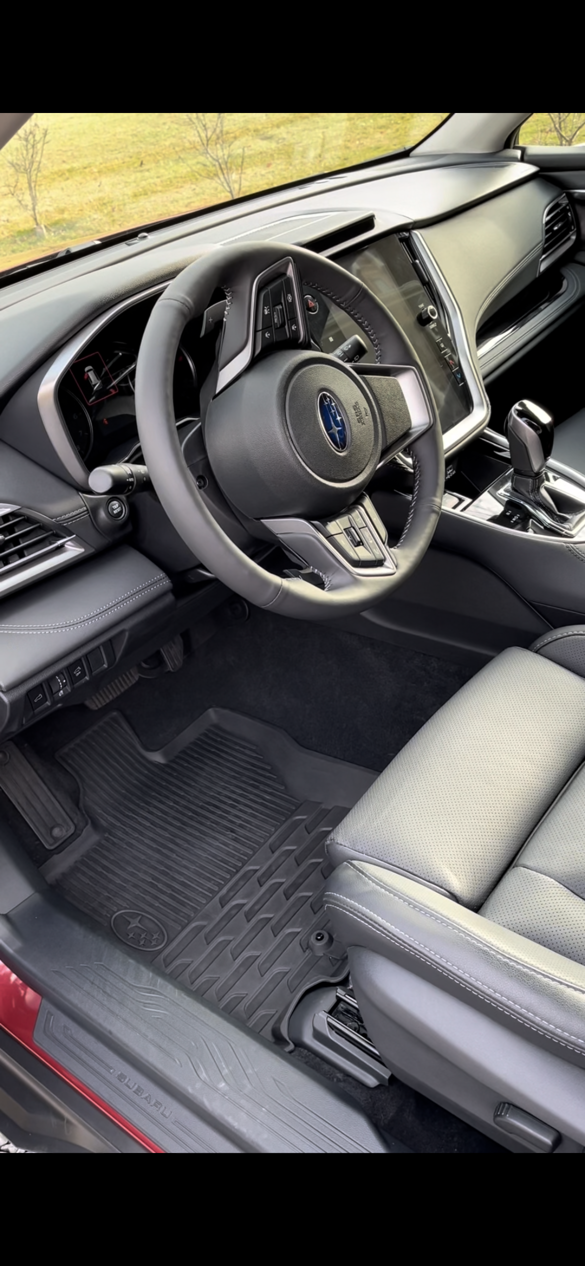 Interior of a Subaru vehicle, showing the steering wheel with Subaru logo, dashboard, center console, gear shift, and front passenger seat with floor mat.