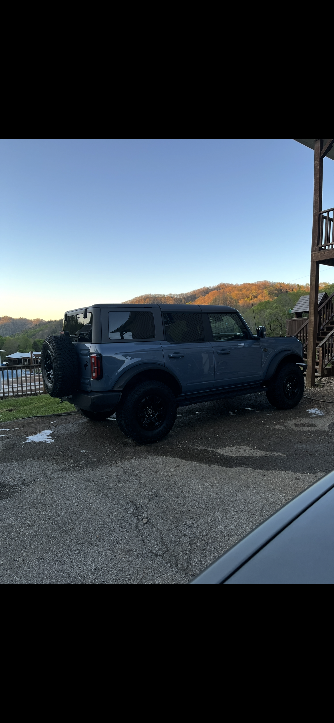 A blue SUV parked next to a wooden building with a mountain and trees in the background during sunset.