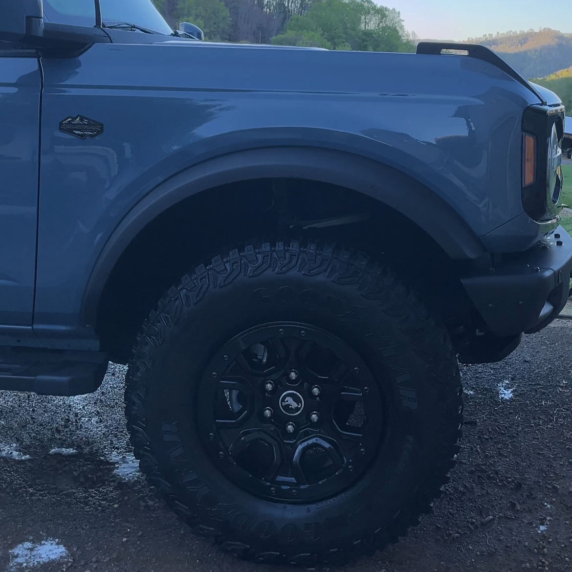 Close-up of a blue pickup truck's front left side, showing the tire, fender, and part of the the headlight, with a scenic background of hills and trees.